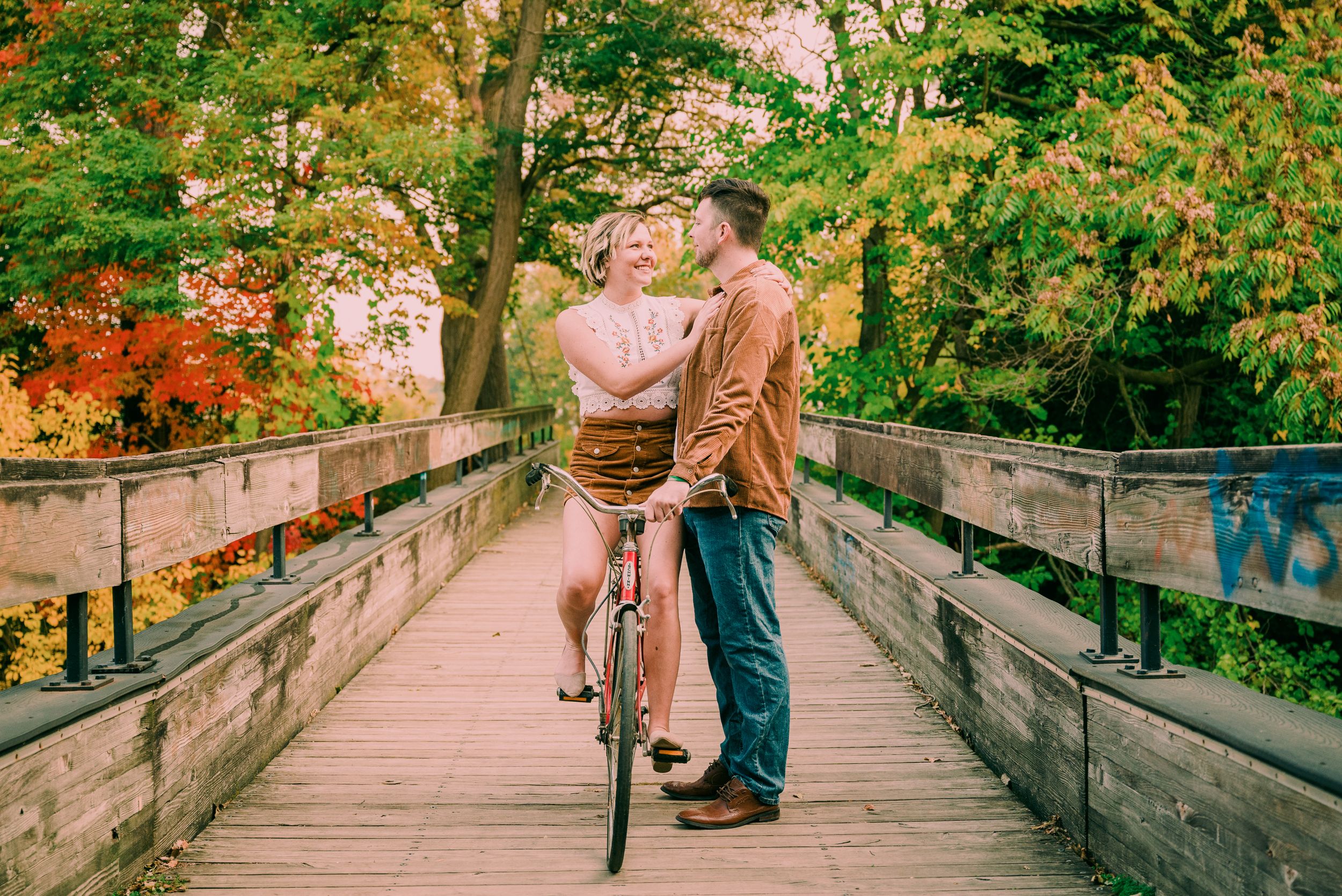 Couple sharing a romantic moment on a wooden bridge with a bicycle amid fall foliage.