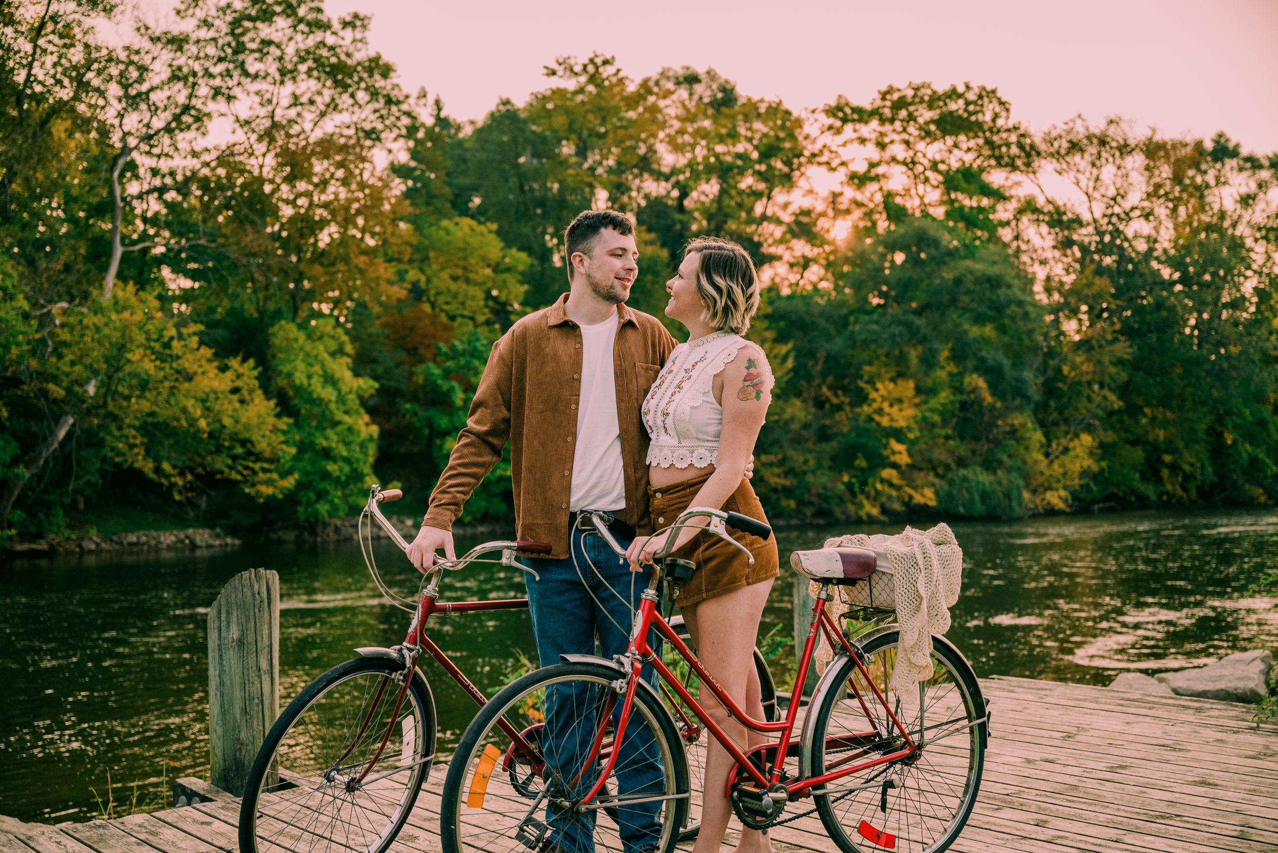 Romantic couple standing with vintage bicycles near a scenic river at golden hour sunset.