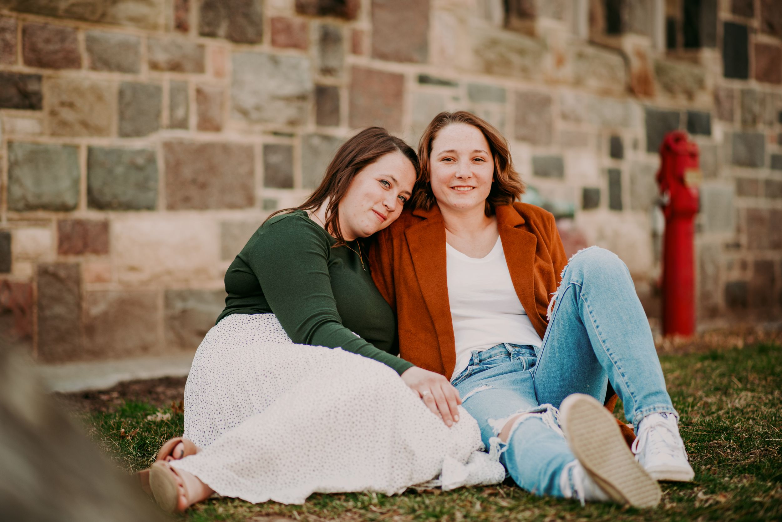 Two women sitting together on grass leaning on each other smiling in front of a stone wall.