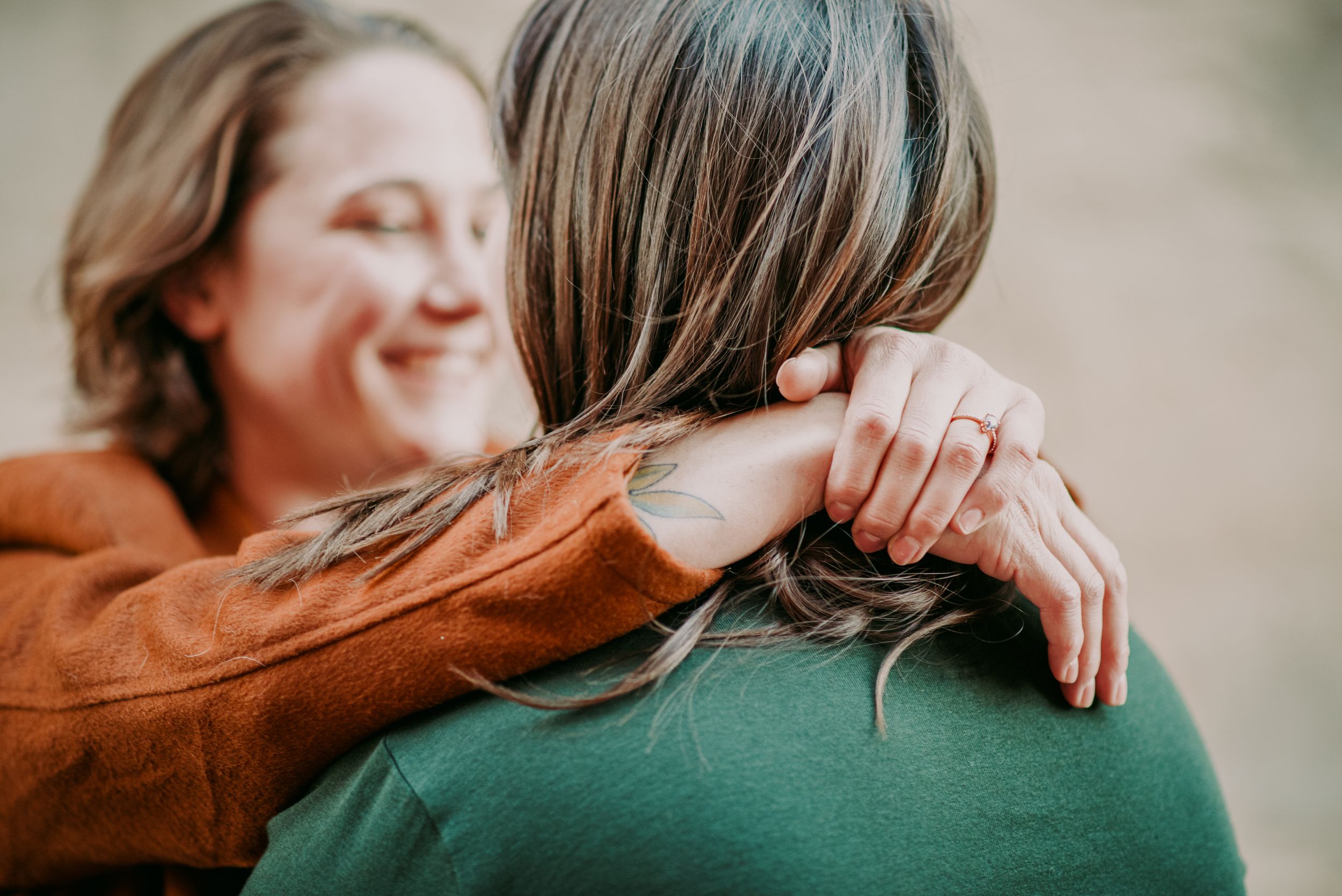 Woman in orange jacket hugging another person wearing green top showing engagement ring.