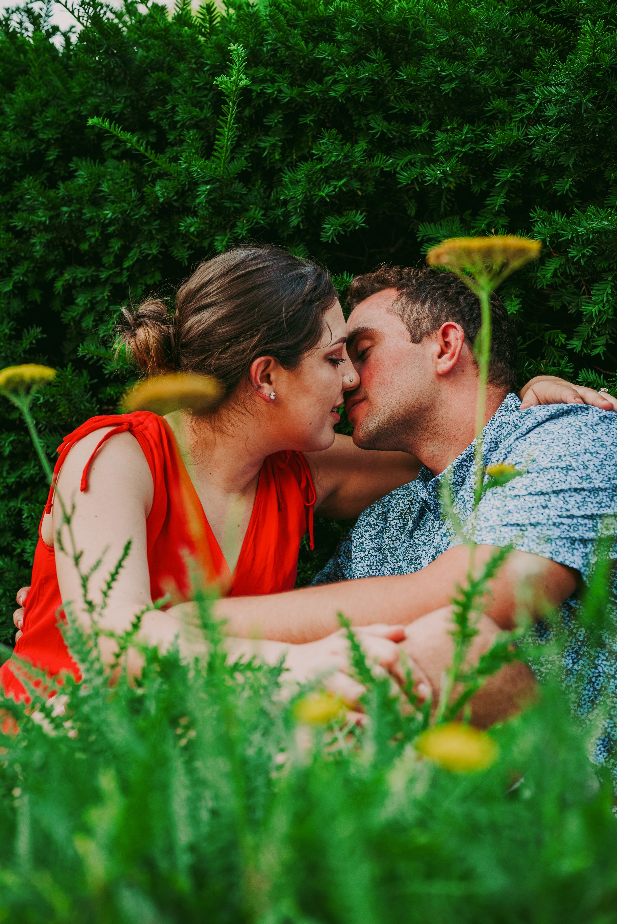 Couple kissing among wildflowers and lush green bushes in a romantic outdoor garden setting.