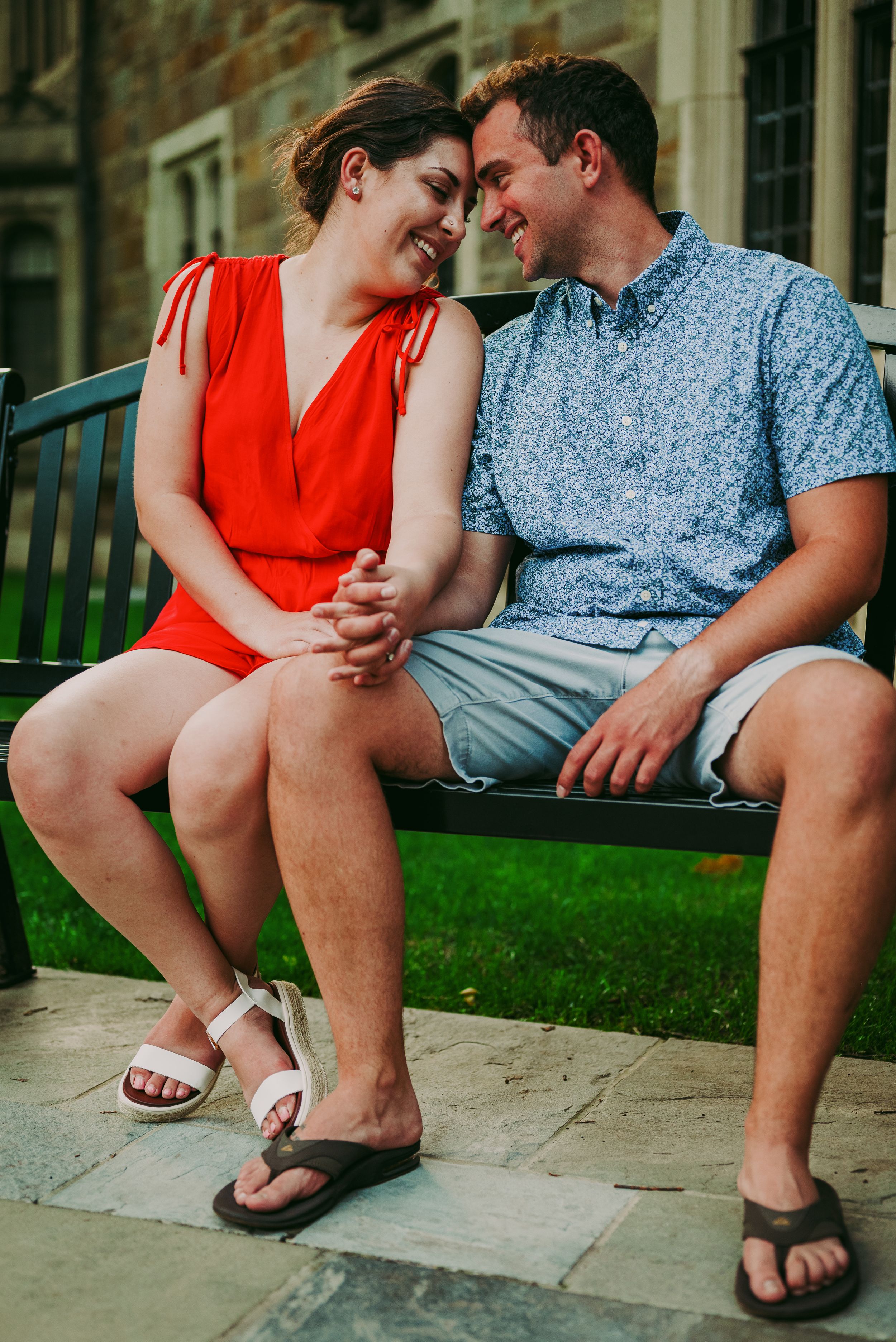 Happy couple sitting on bench touching foreheads and holding hands in front of stone building.