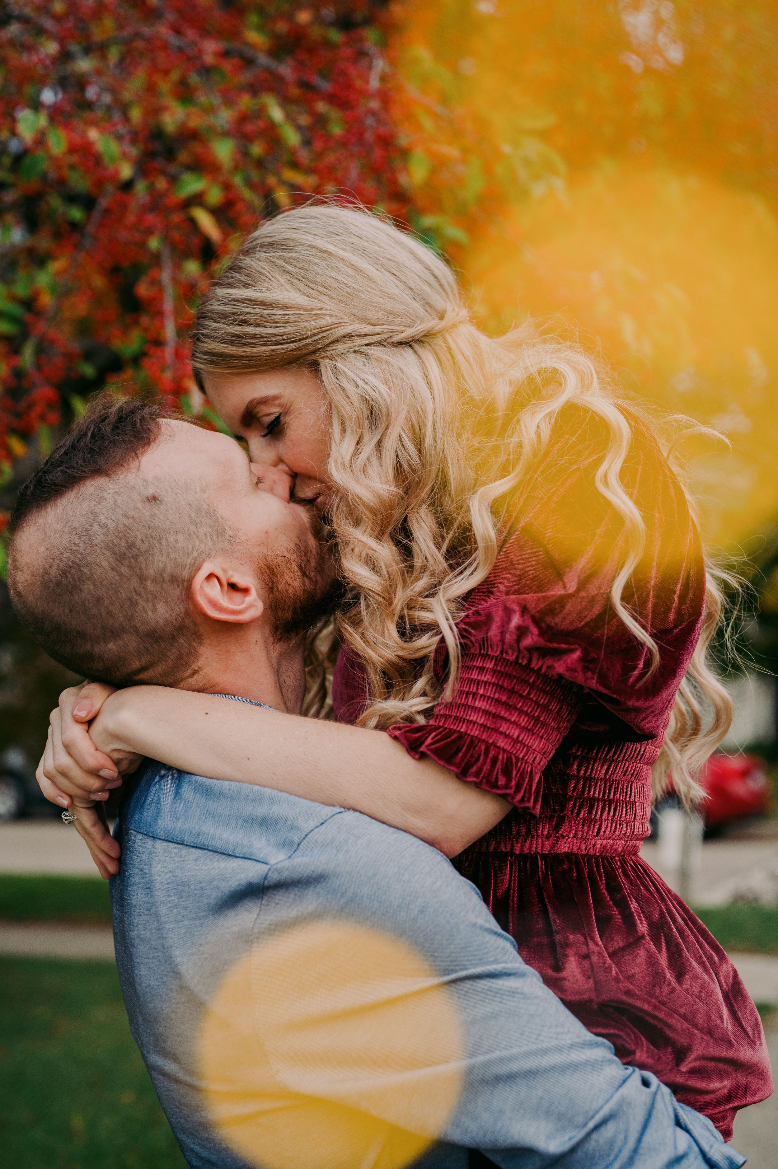 Man lifting woman in burgundy dress for a kiss with vibrant autumn foliage background.