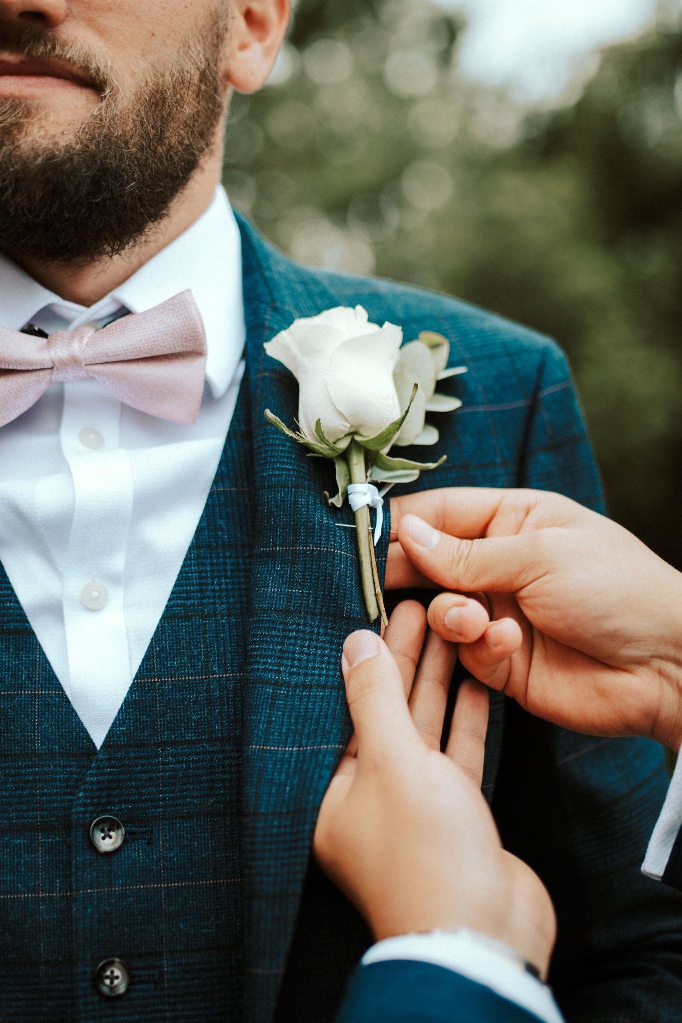 Hands pinning a white rose boutonniere on a navy checkered suit with pink bow tie. Saltmarshe Hall
