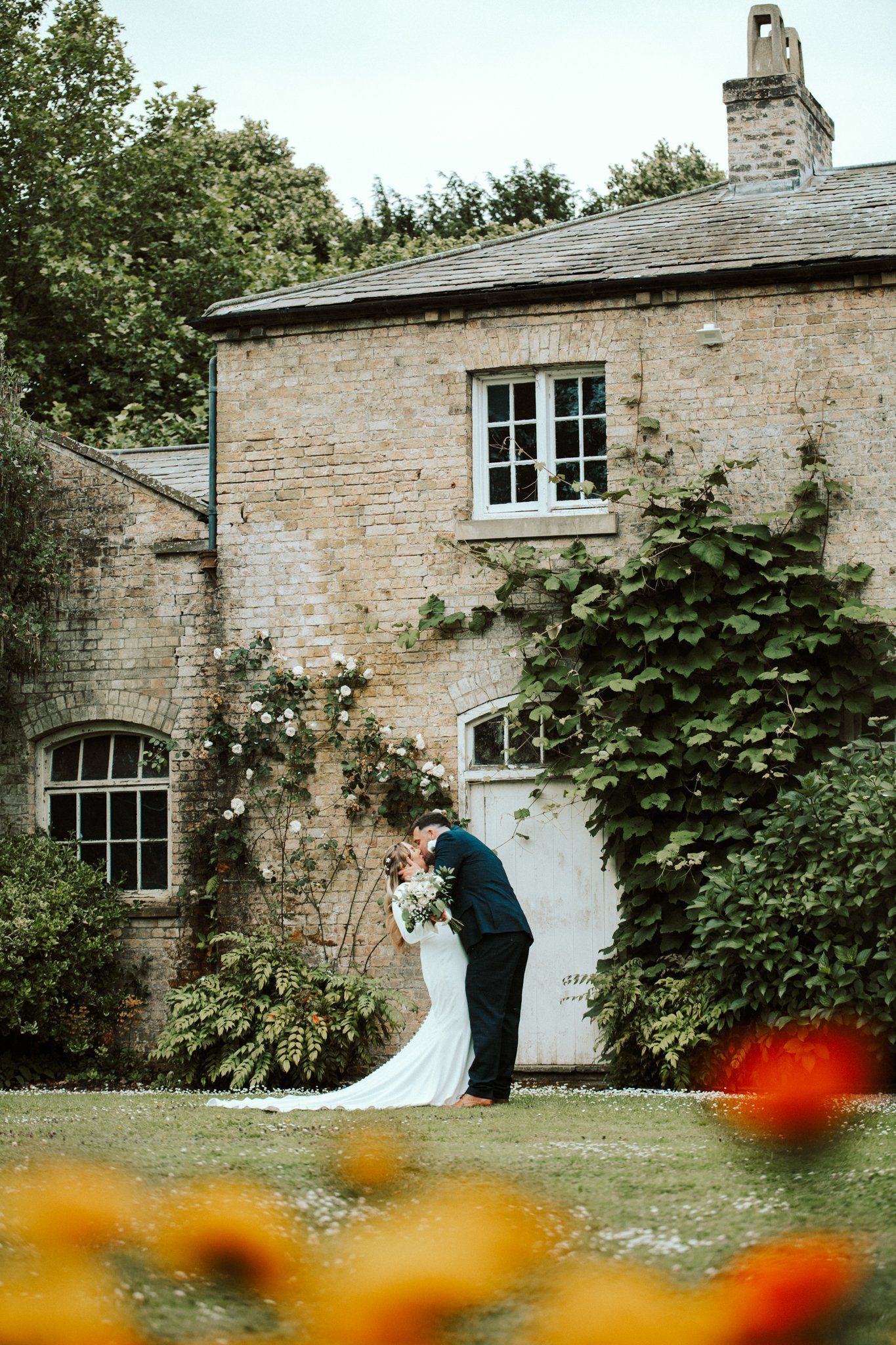 A wedding couple embraces in front of an ivy-covered cottage with orange blooms in view. Saltmarshe Hall