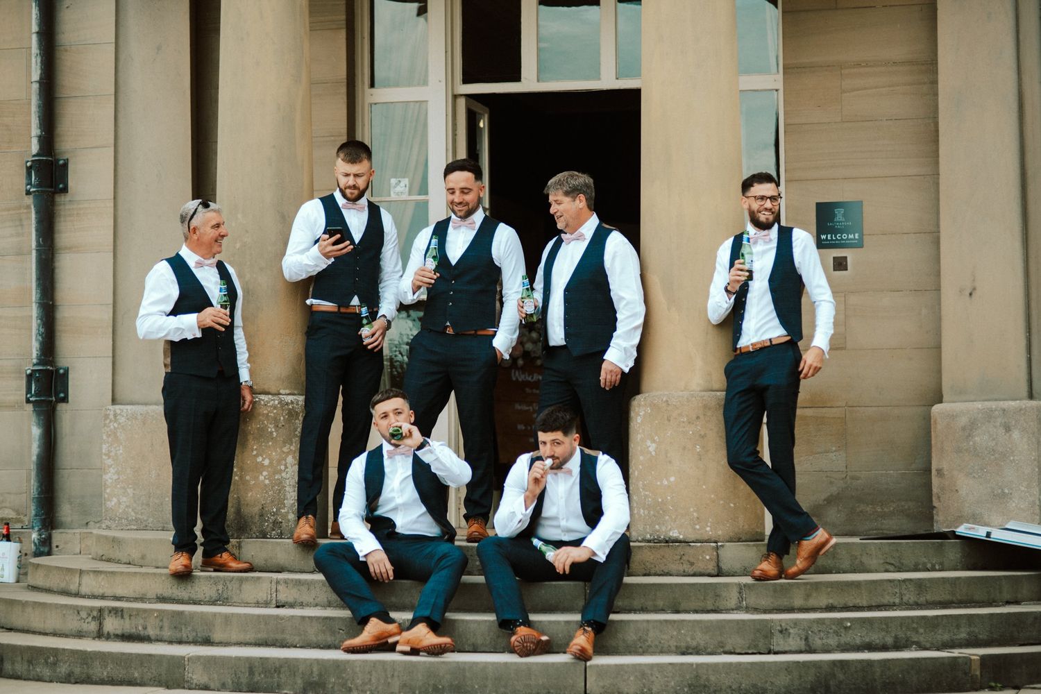 Groomsmen in matching vests and formal wear sit casually on stone steps outside. Saltmarshe Hall