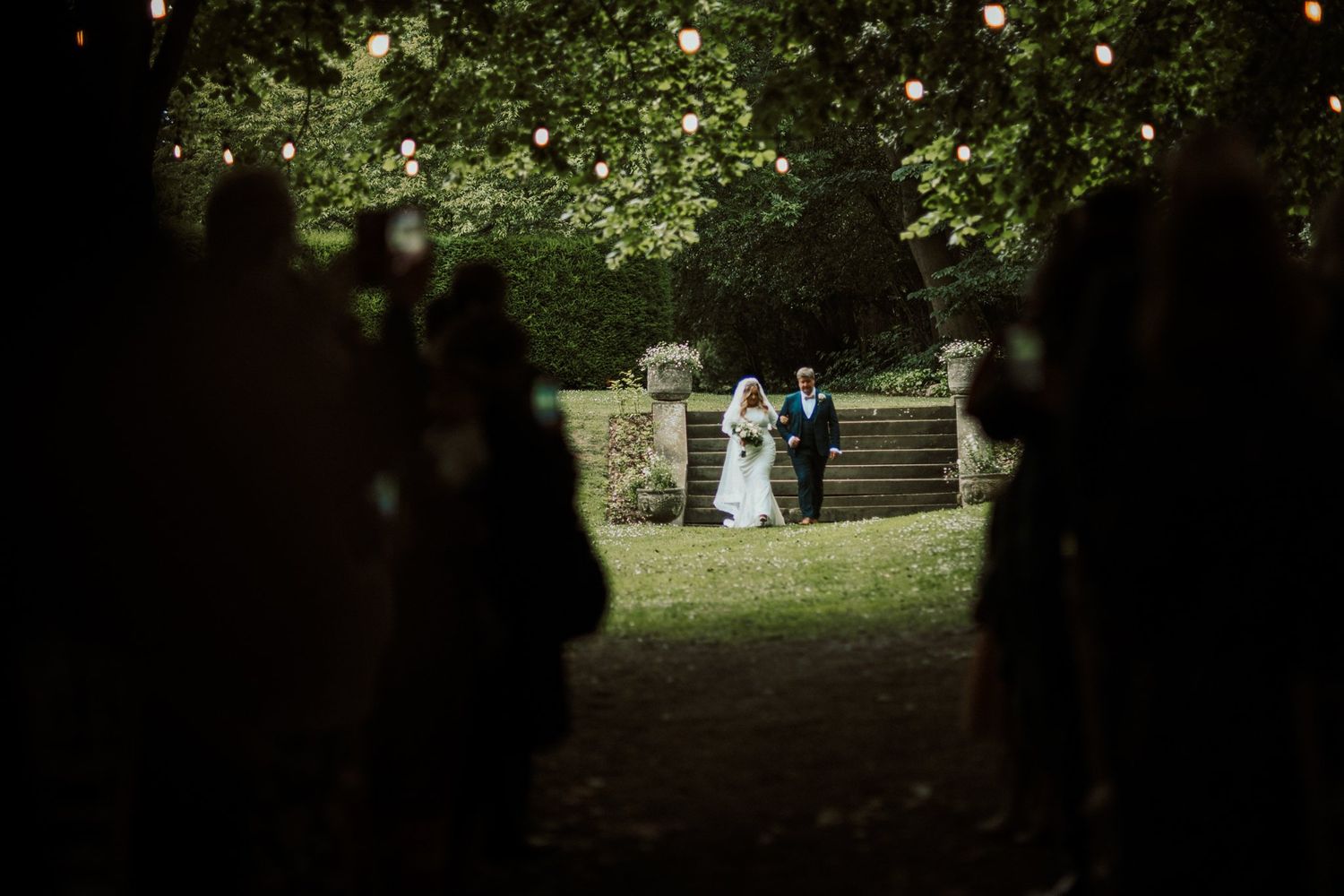 Atmospheric view of a wedding ceremony through trees with string lights glowing. Saltmarshe Hall