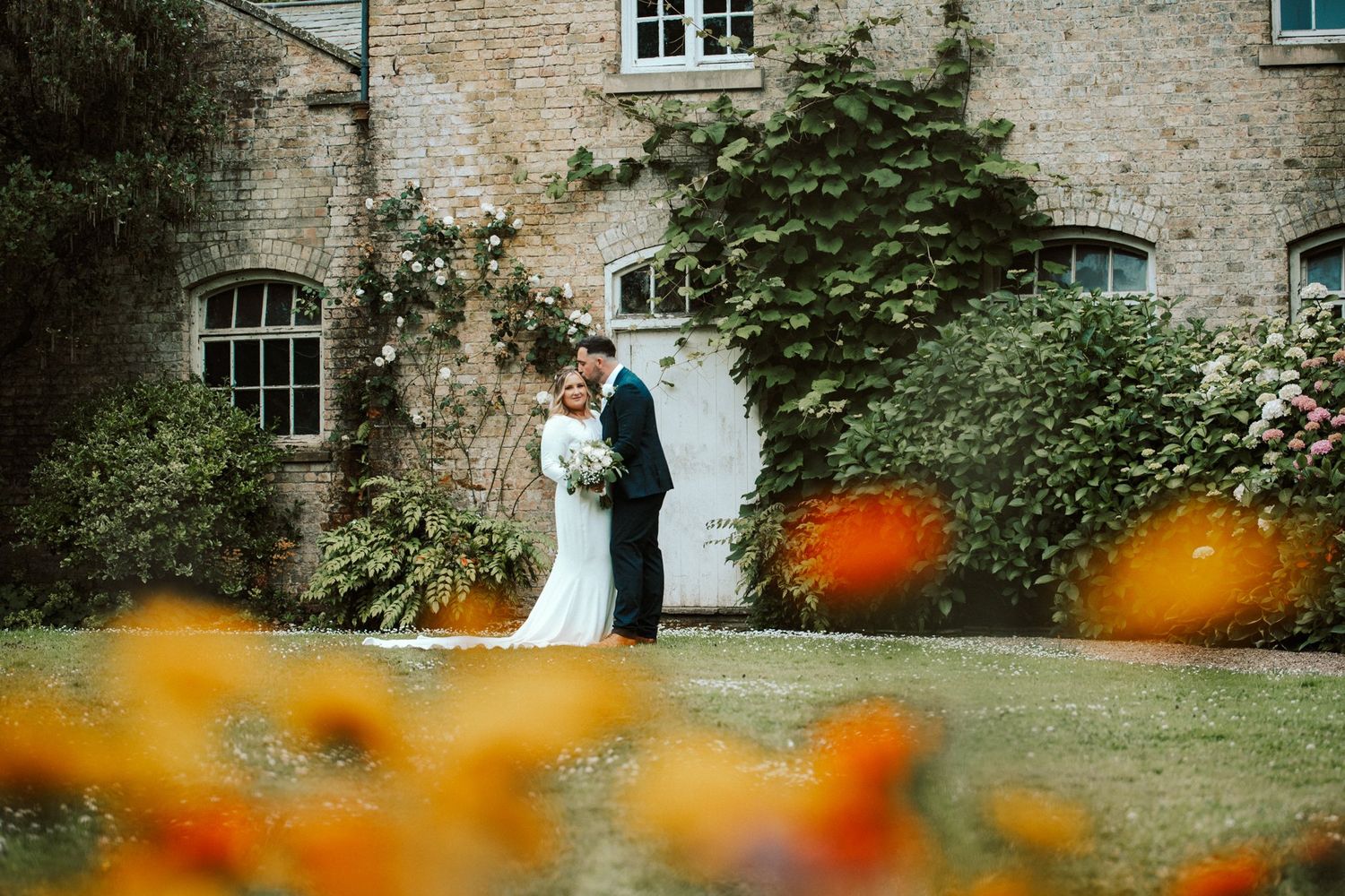 Newlyweds pose by a rustic stone building with orange flowers in the foreground. Saltmarshe Hall