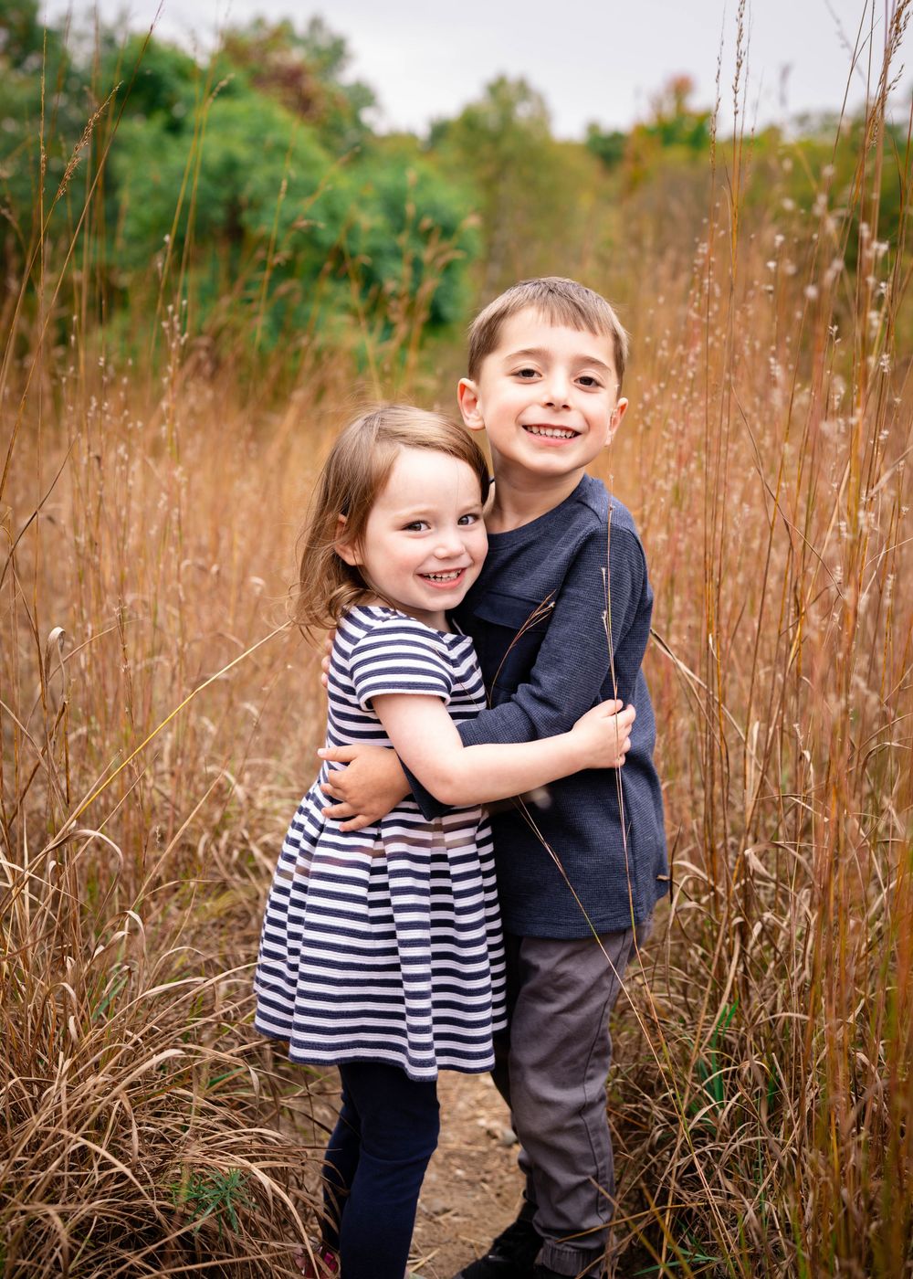 A young brother and sister hugging and smiling together in a tall golden grass field in autumn.