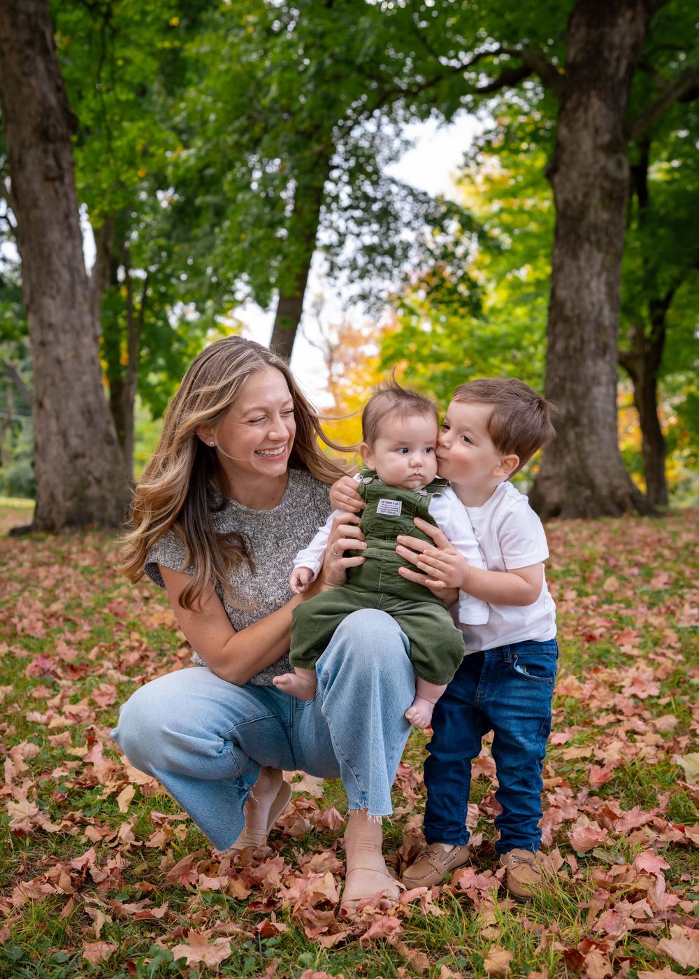 A smiling mother crouches in autumn leaves holding her baby while her toddler son gives the baby a kiss.