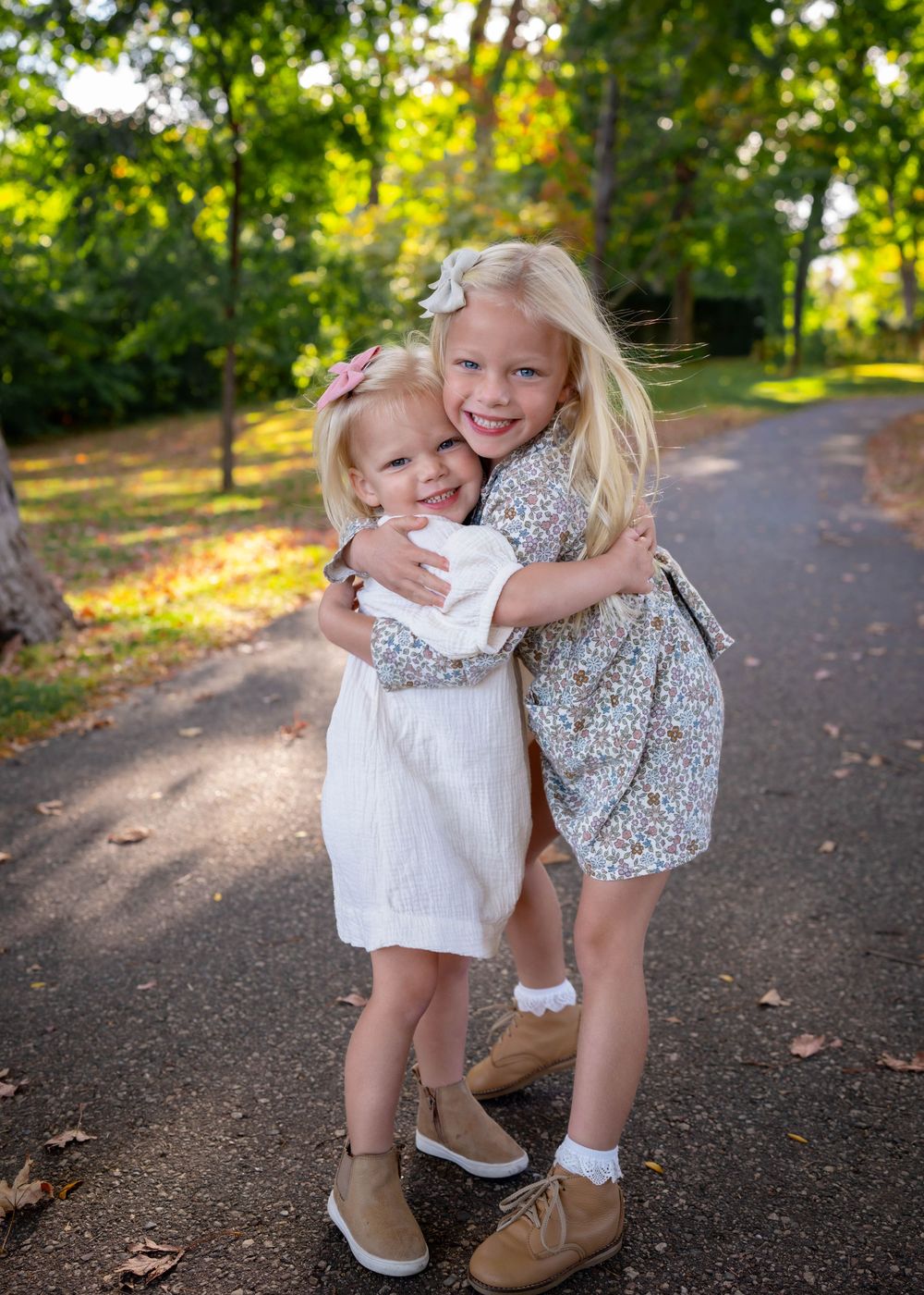 Two blonde sisters hugging and smiling on a tree-lined path during an outdoor fall family photo session.