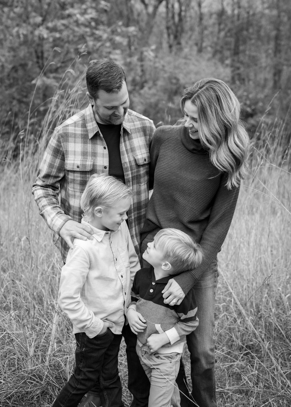 Black and white family portrait of parents with two young sons standing in a golden grass field.