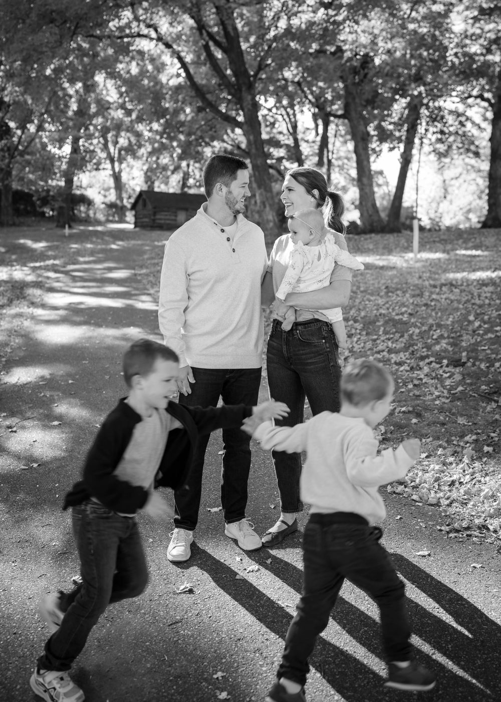 Black and white photo of a couple smiling at each other while two young boys run around them in a park.