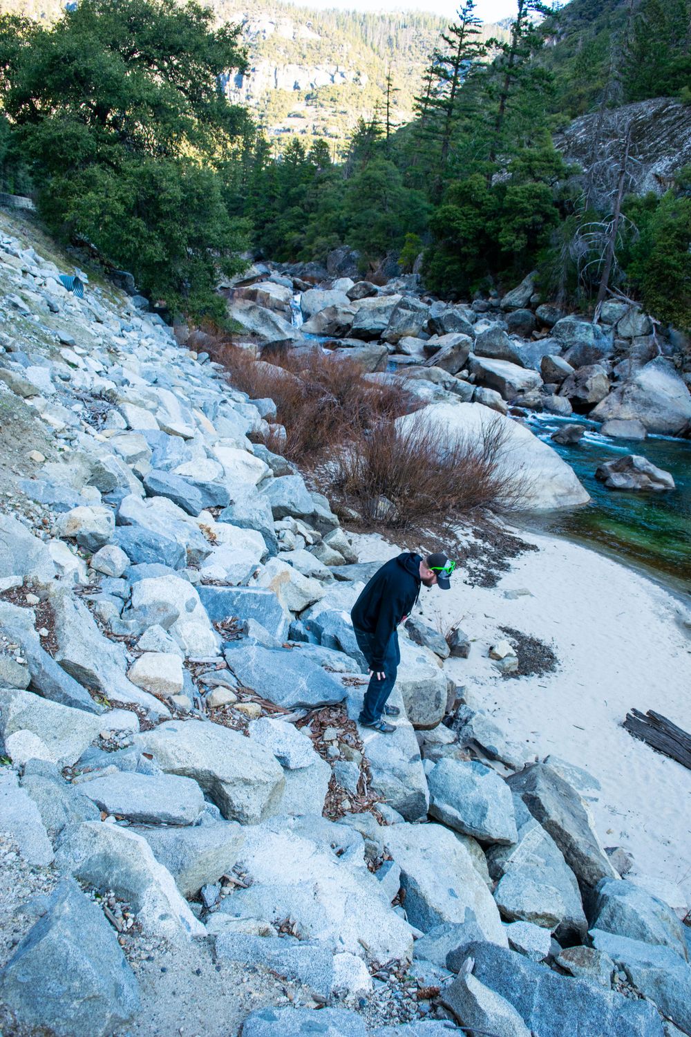 Explorer traverses rocky slope near turquoise mountain stream in winter landscape.