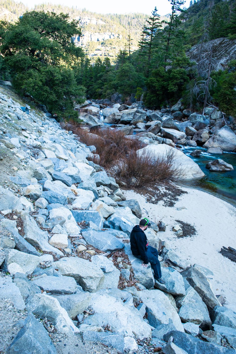 Hiker explores rocky riverbank with turquoise water and snow patches in mountain valley.