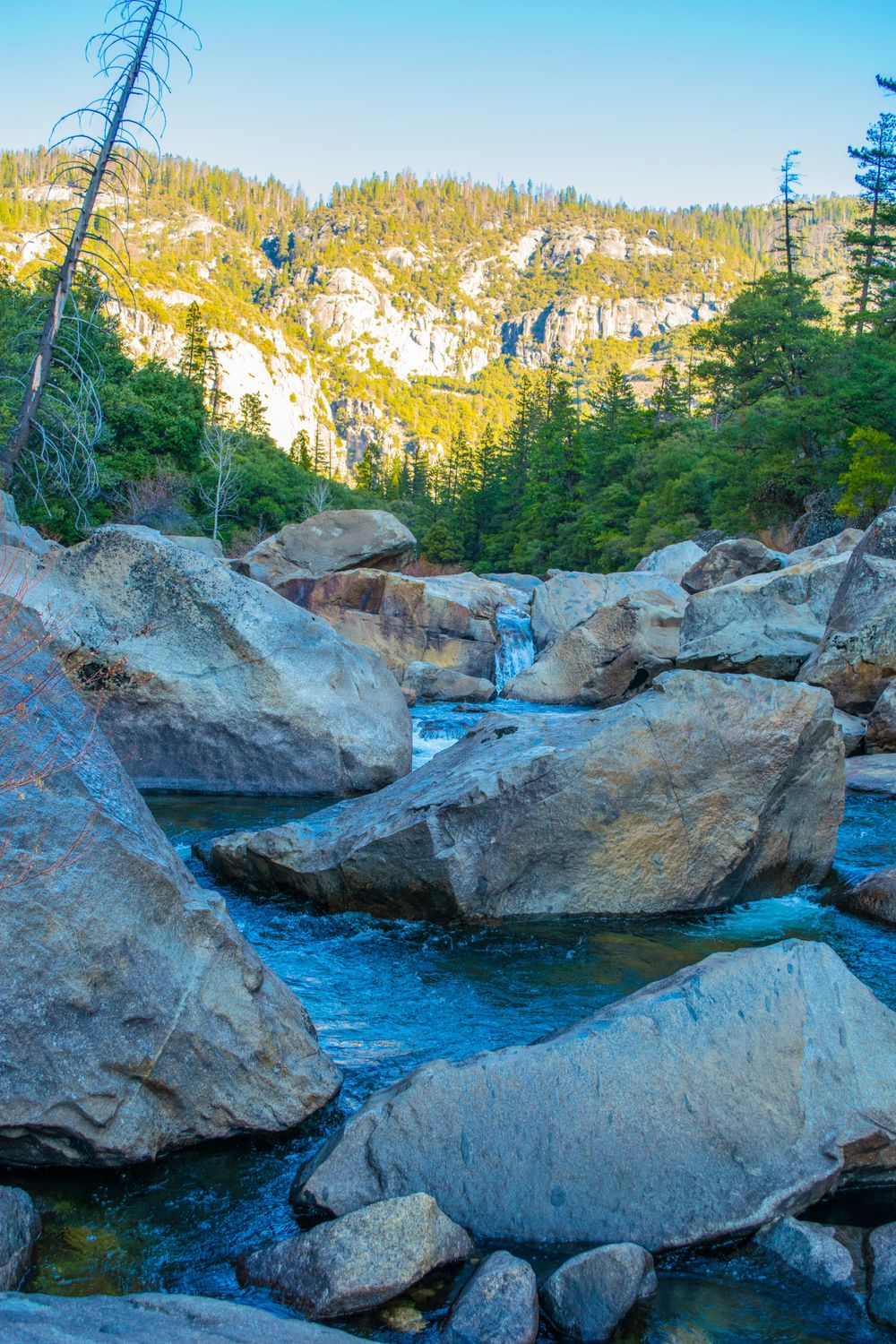 Crystal clear mountain stream flows between massive boulders with snow-capped peaks in background.