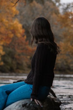 a girl sitting on a tree truck in a shallow part of a river with fall leaves showing