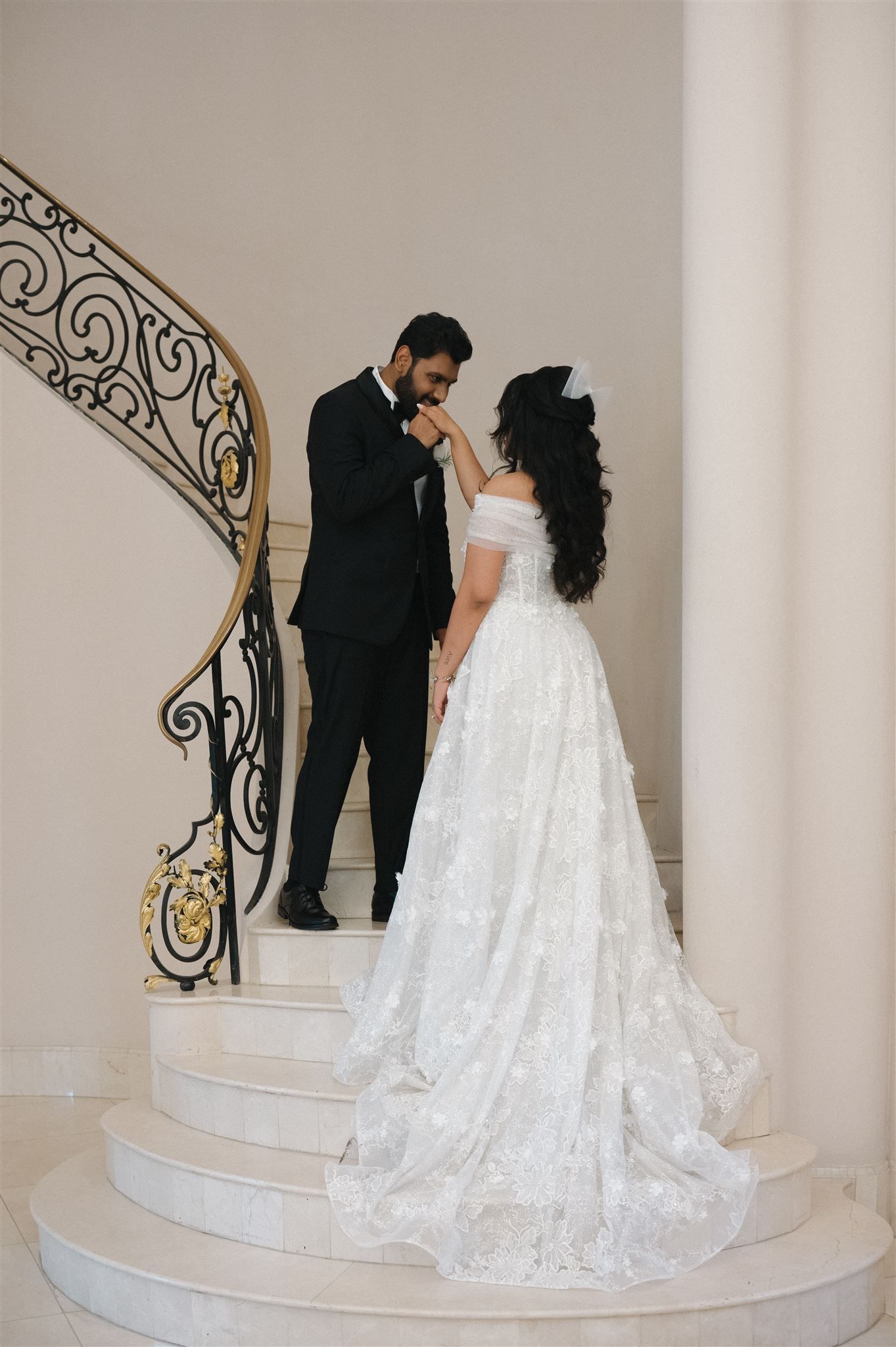 Elegant couple in formal wedding attire stands together on white marble staircase with ornate wrought iron railing.