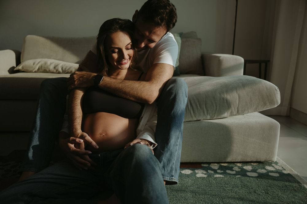 Couple sharing tender moment on floor, pregnant woman cradled by partner, soft natural light in cozy living room.