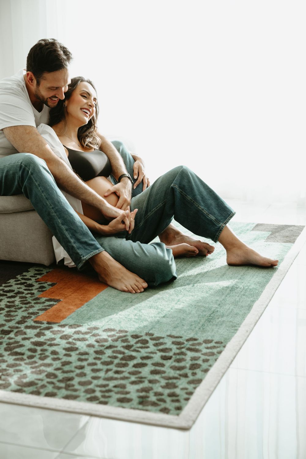 A happy couple sits on a cozy rug, leaning against a sofa, smiling and embracing in a bright living room.