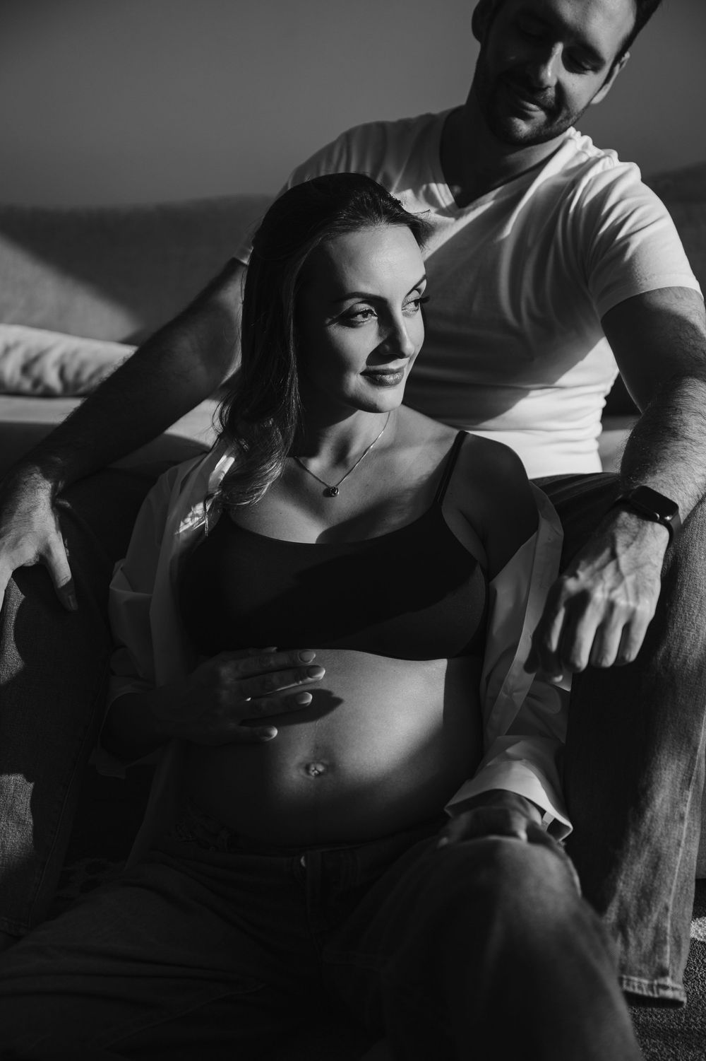 Black and white photo of a smiling pregnant woman sitting with her partner, gently cradling her baby bump.