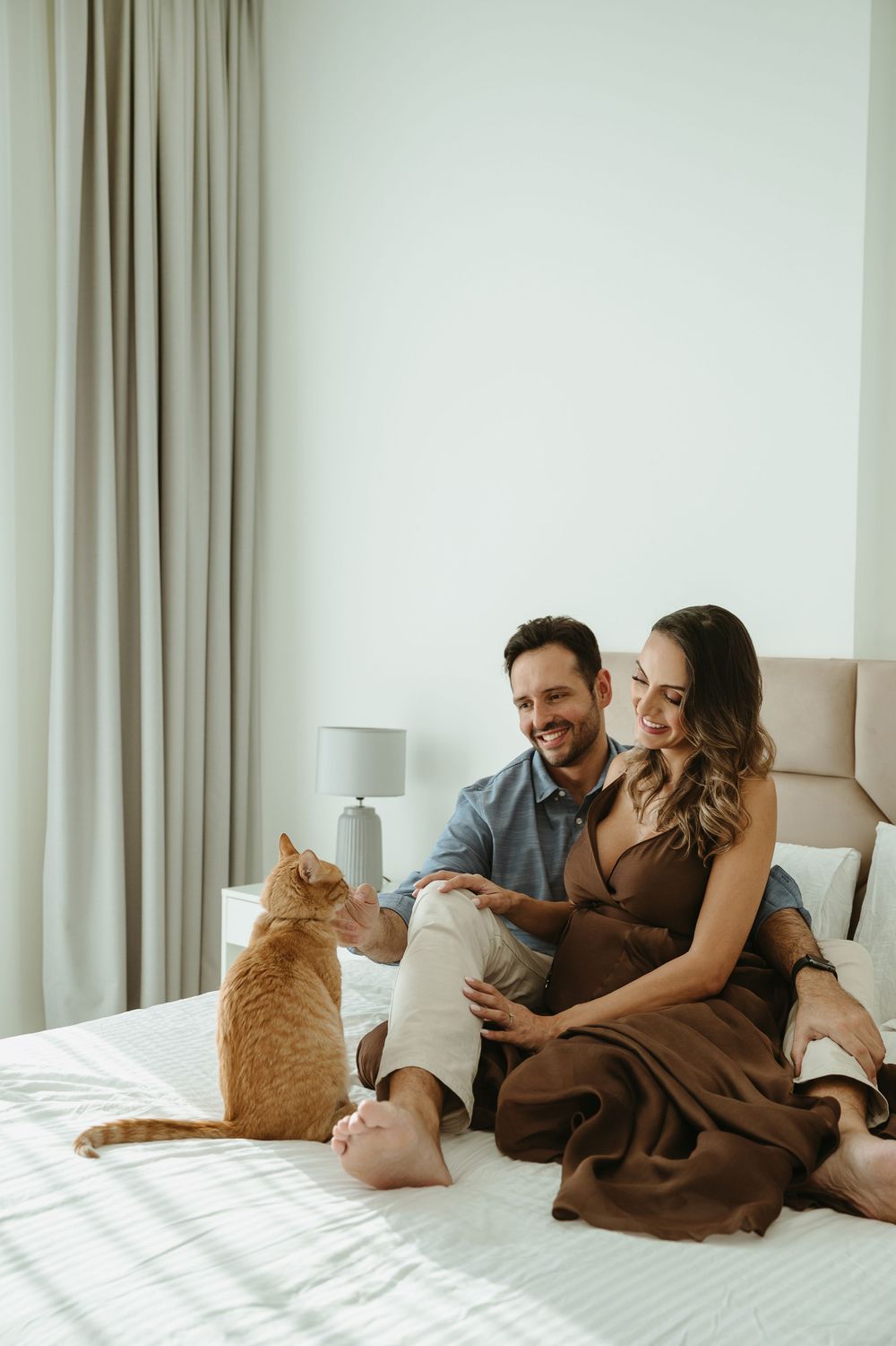 A smiling couple sits on a white bed interacting with an orange tabby cat in a bright, modern bedroom.