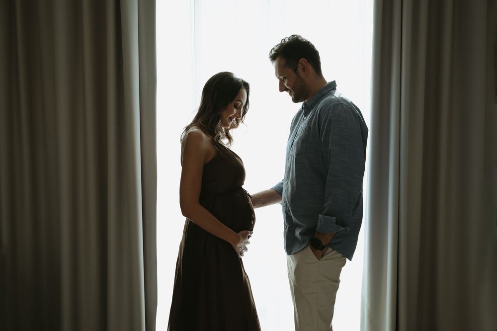 Pregnant woman in dark dress with partner standing by curtained window in intimate maternity photo shoot.