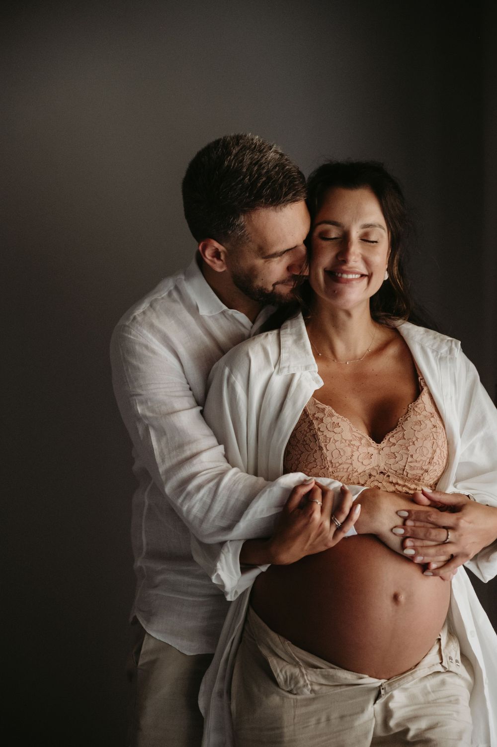 A man tenderly embraces a smiling pregnant woman from behind, both wearing white, against a gray background.