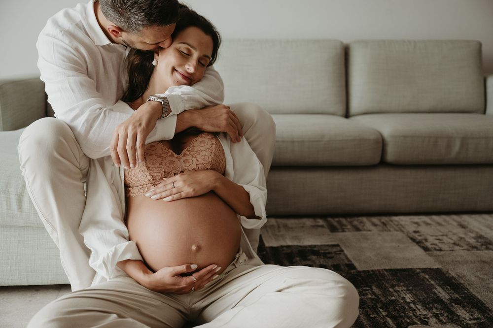 A couple sitting on the floor, the man embracing his pregnant partner as she cradles her baby bump near a gray sofa.