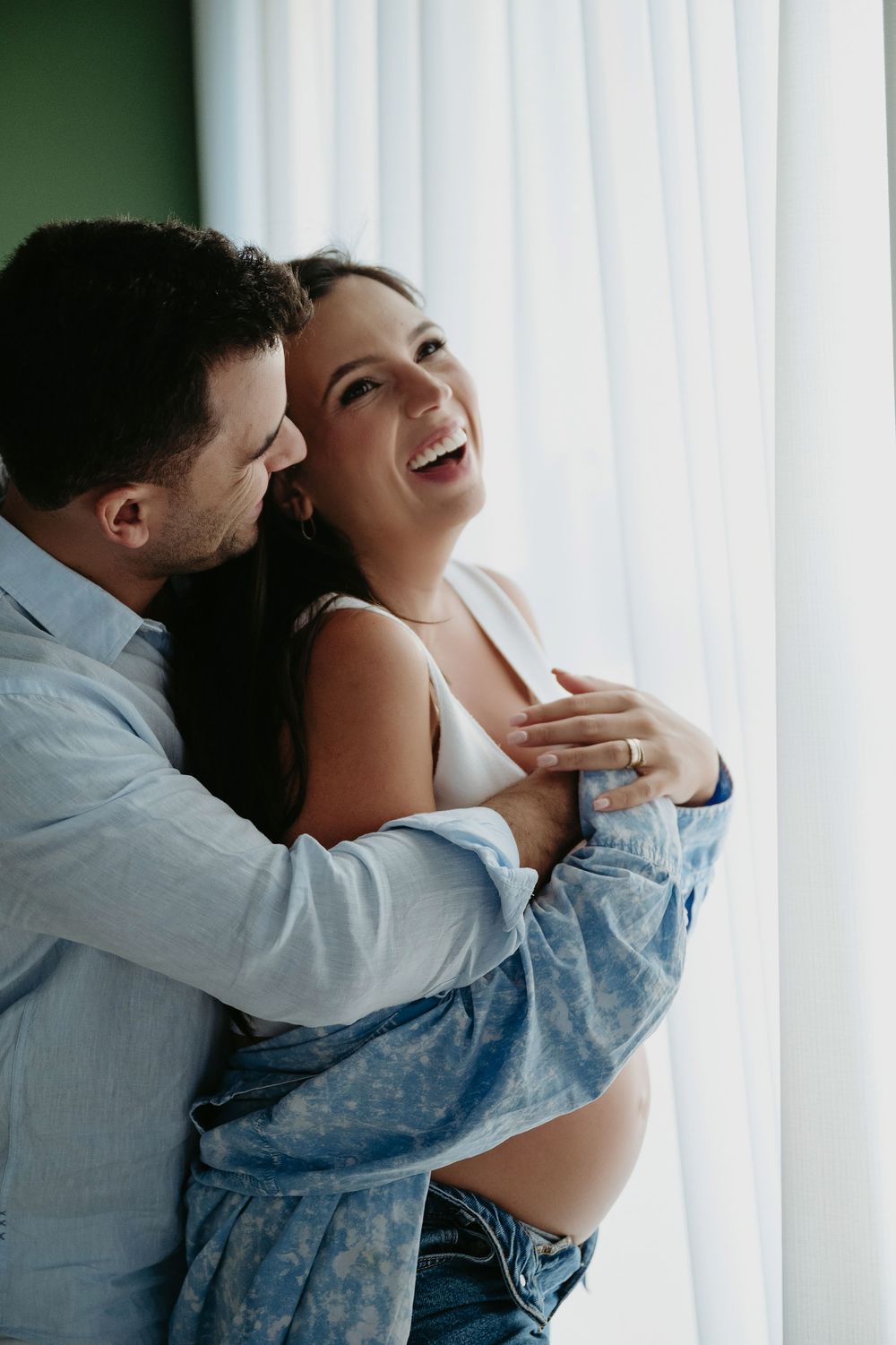 A happy pregnant woman laughs as her partner embraces her from behind near a bright window.
