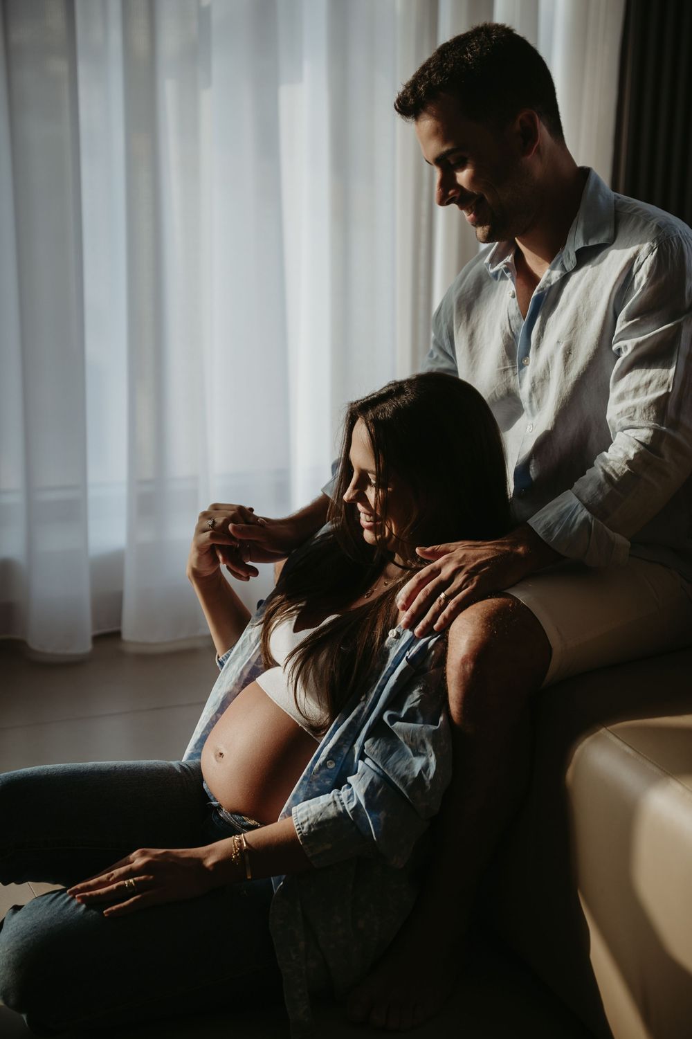 Couple sharing a tender moment, pregnant woman sitting on floor leaning against sofa as partner stands behind her.