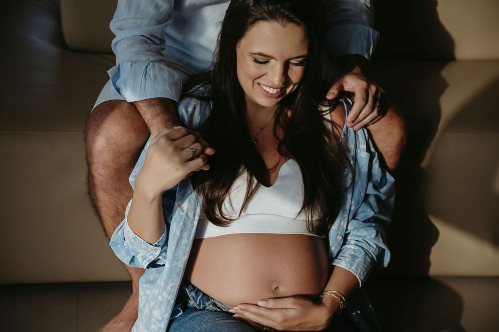 Smiling pregnant woman in white crop top cradling baby bump while partner embraces her from behind on couch.