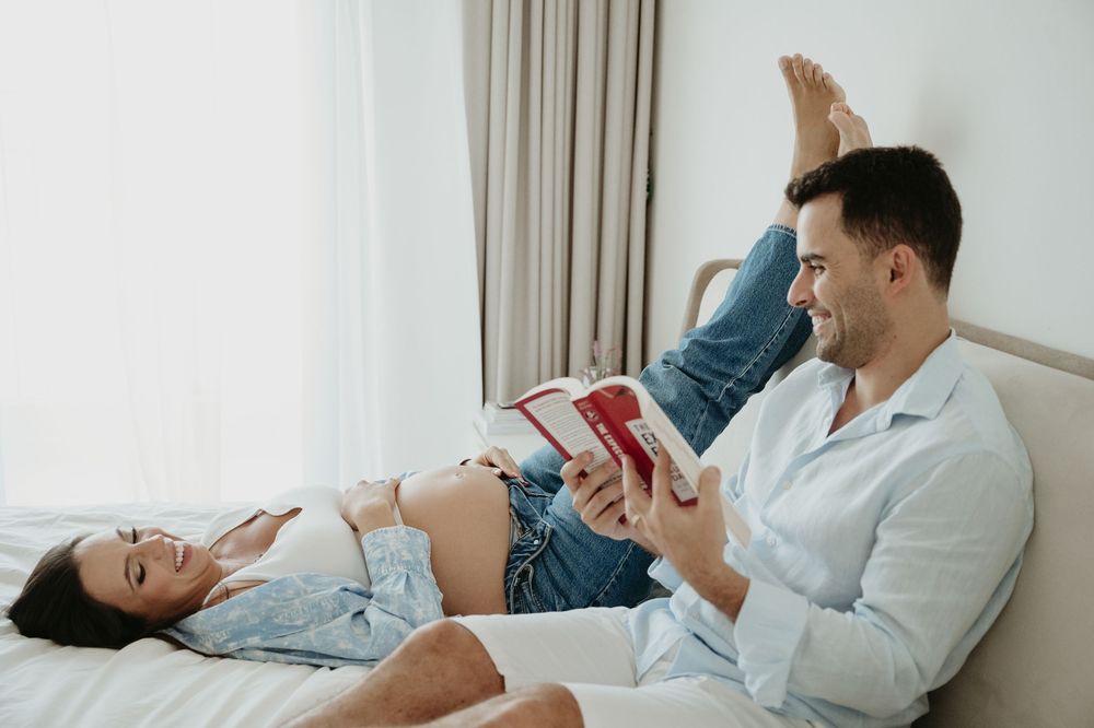 Smiling couple relaxing on bed, pregnant woman lying down while man reads red book to her belly.