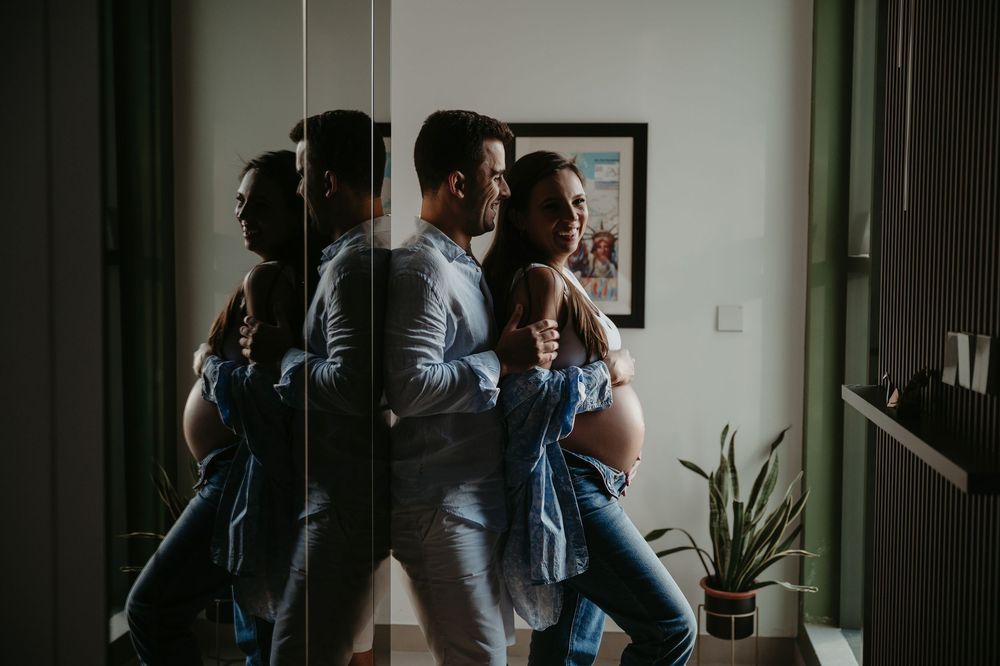 Couple leaning against mirror wall, woman with baby bump, soft natural light in cozy room with plant.