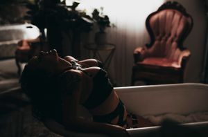 Woman in black lingerie, leaning back in a vintage bathtub, with soft lighting casting shadows over her seductive pose.
