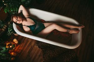 Woman in green lingerie, lying in a vintage bathtub surrounded by plants and soft candlelight, exuding a relaxed and sensual vibe.