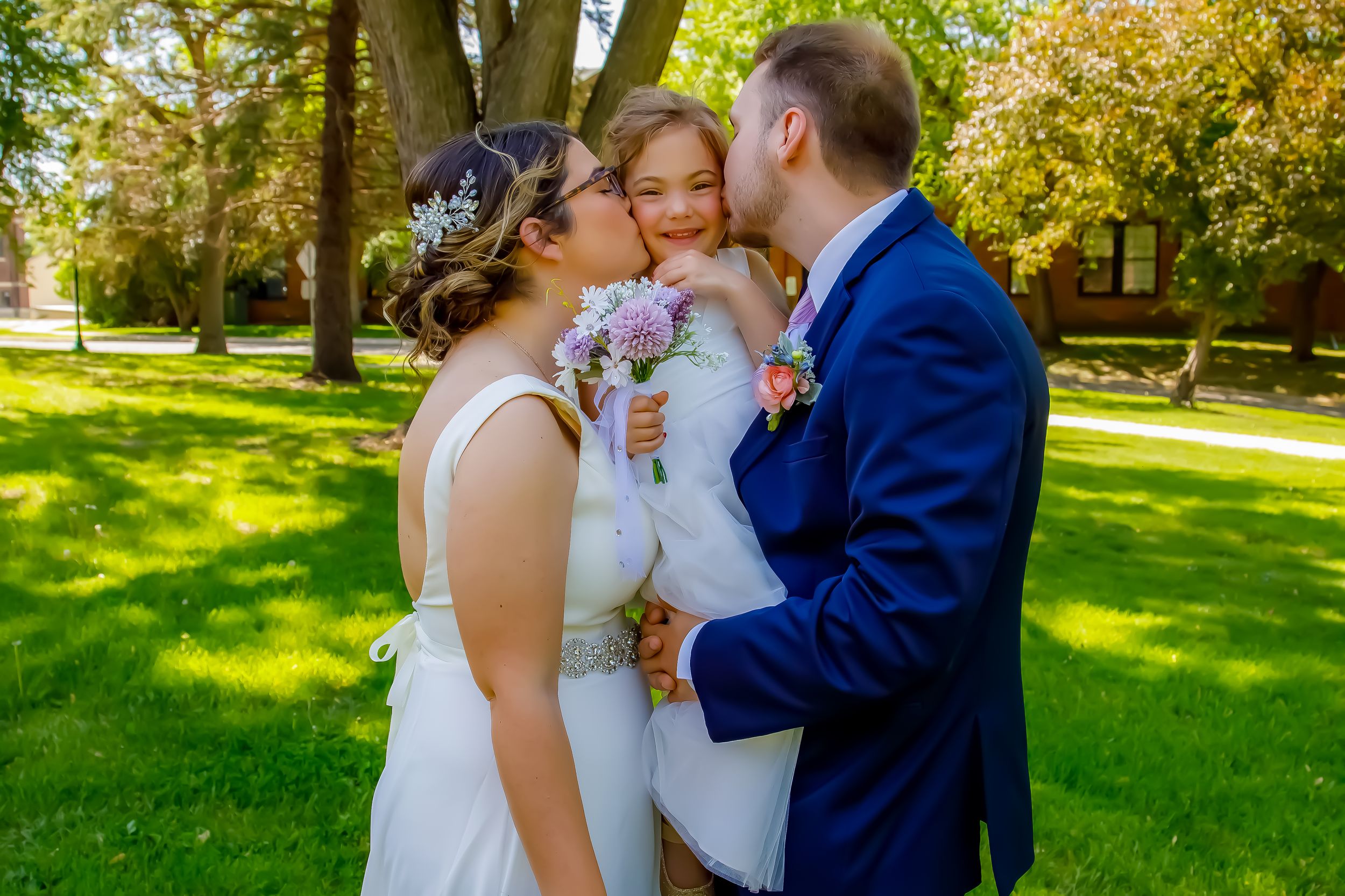 A joyful wedding moment captured outdoors with three people embracing on sunny grass.