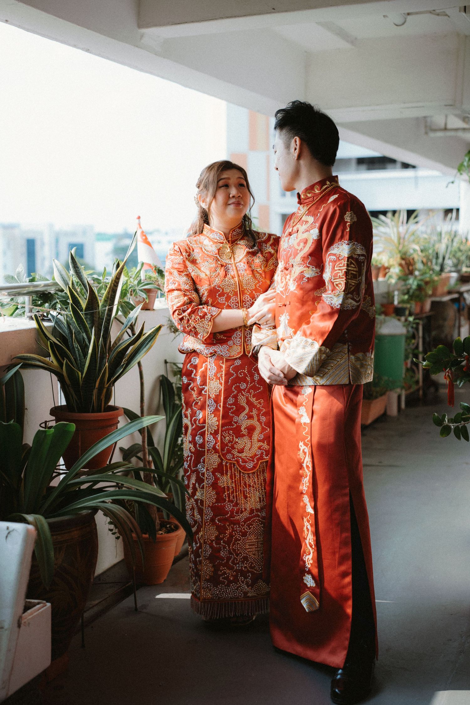 A couple in matching red and gold traditional Chinese attire poses on a balcony with plants.