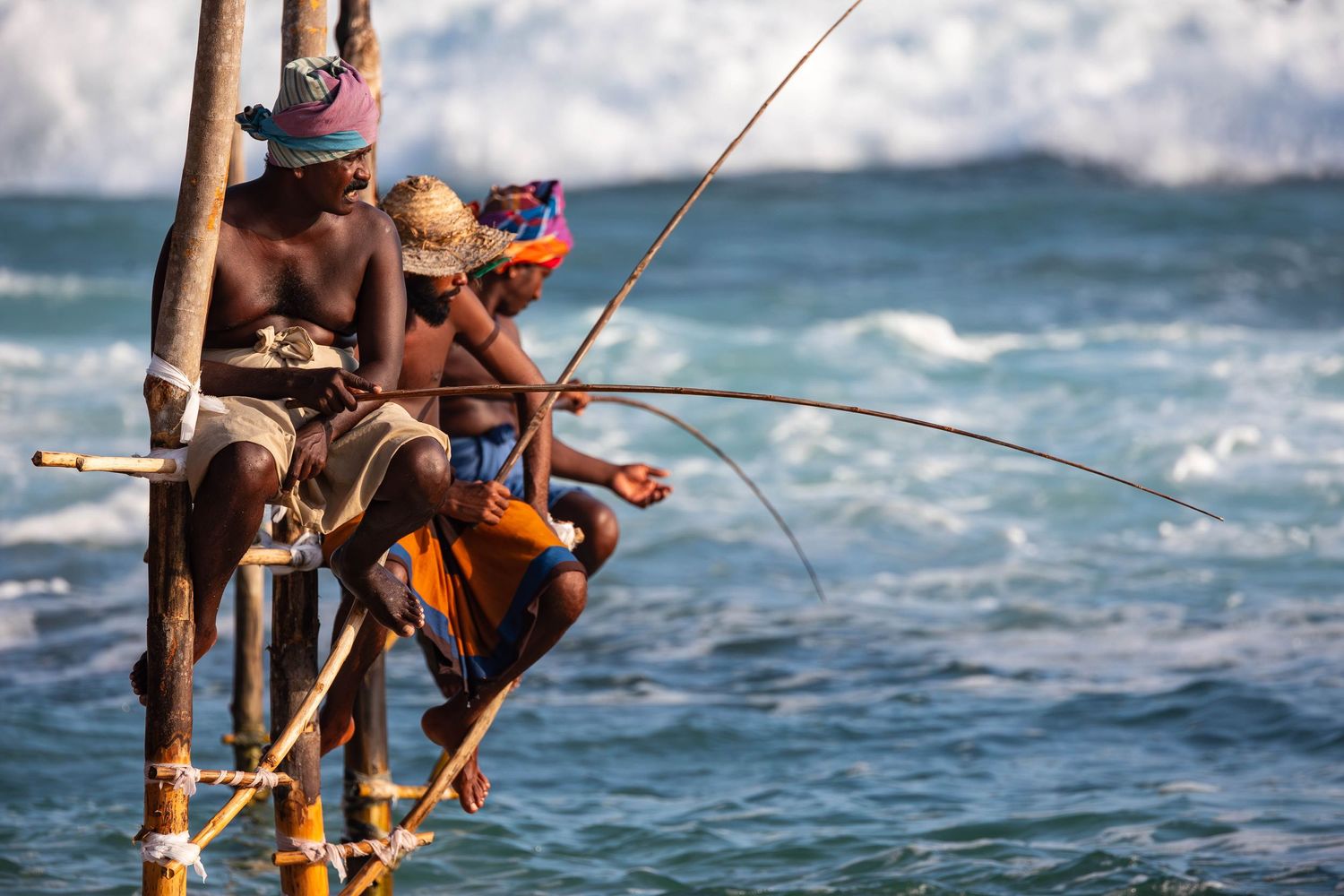 Traditional stilt fishermen casting lines into ocean waves off tropical coast.