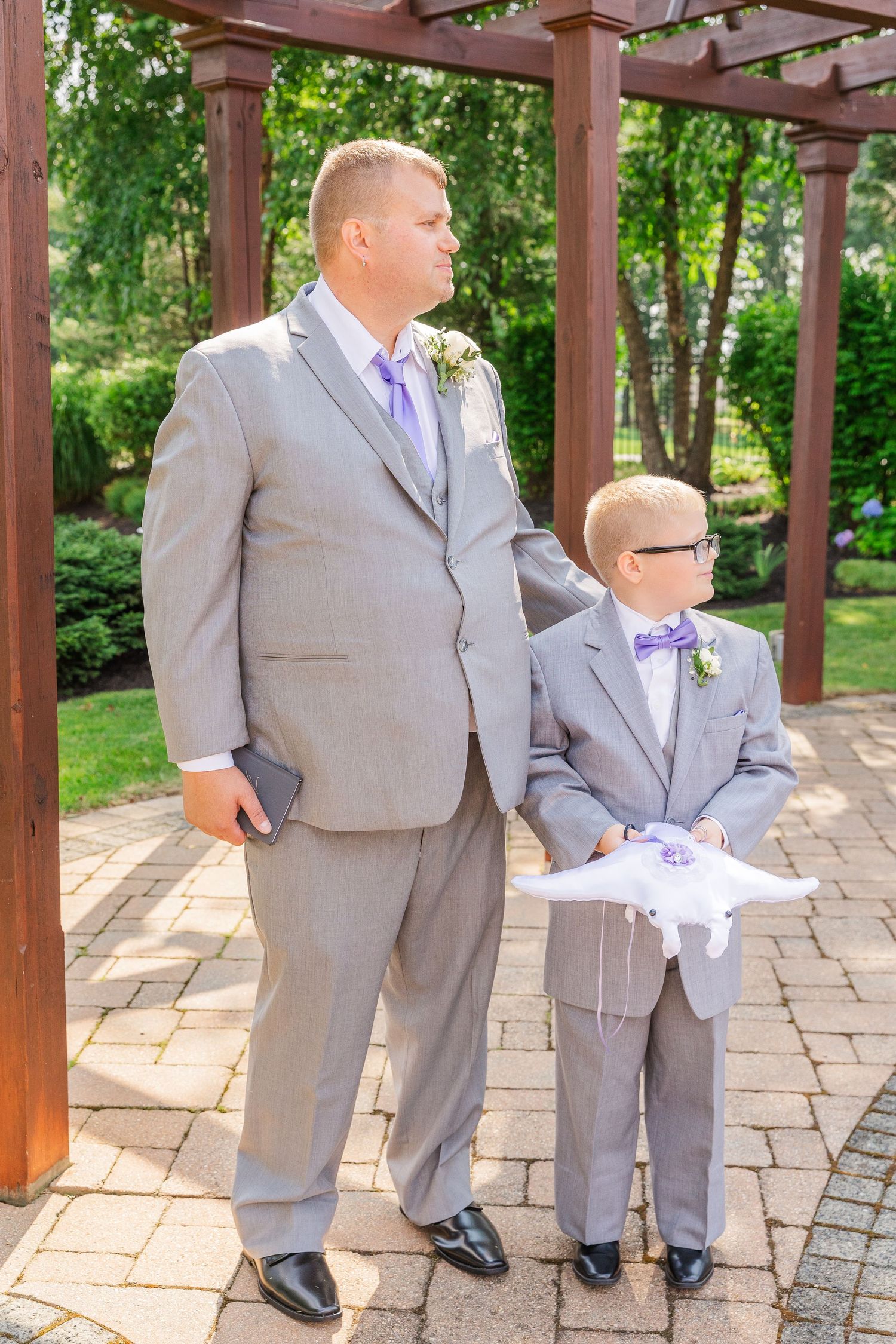 Man and young boy in matching gray suits standing under wooden pergola.