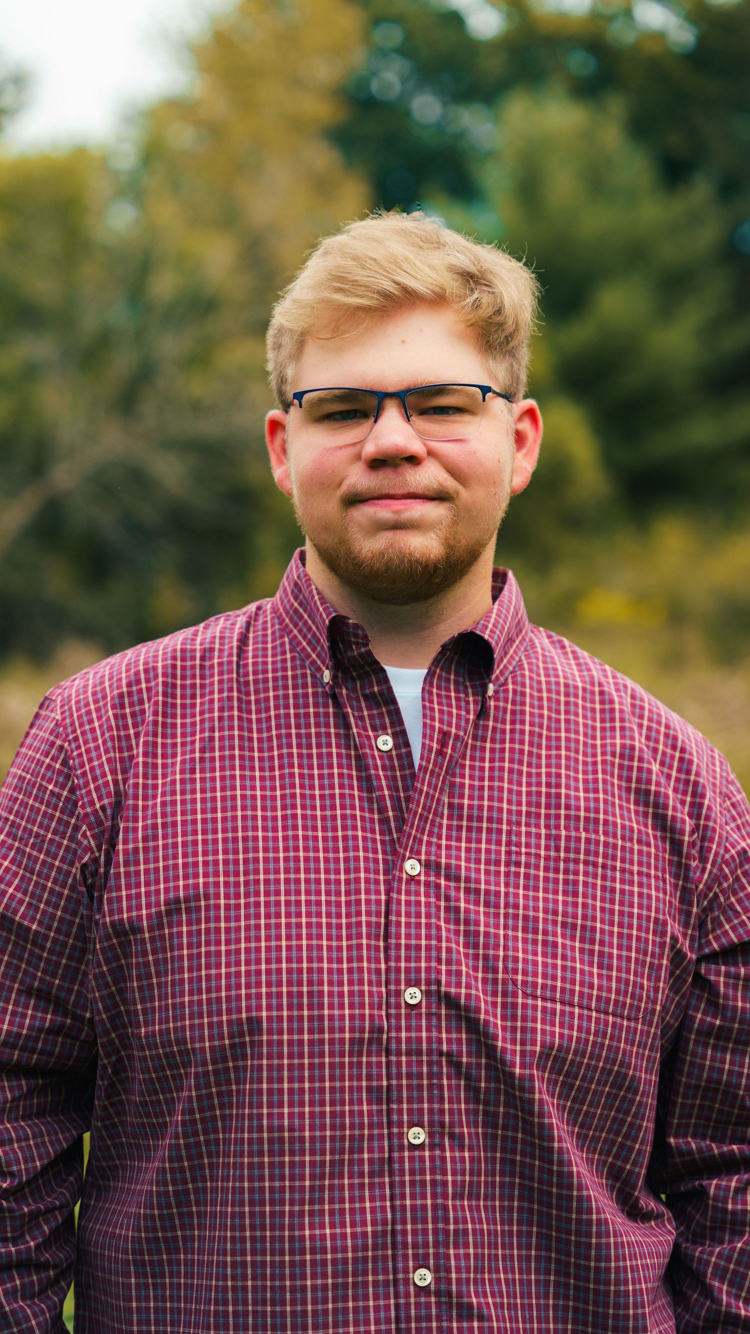 A photographer wearing glasses and a red plaid shirt holds a professional camera while shooting outdoors in a natural setting.