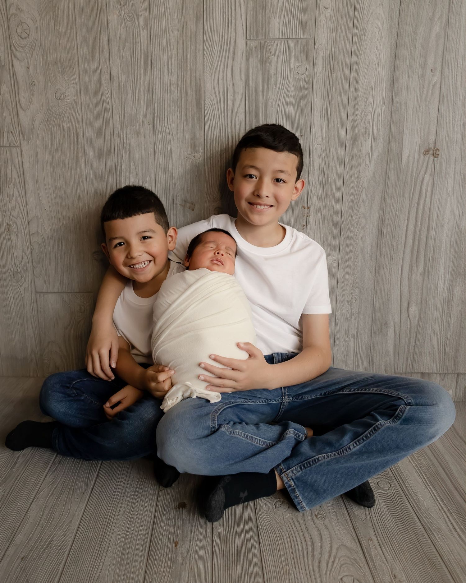 Three siblings pose together against wooden wall background during newborn photoshoot.