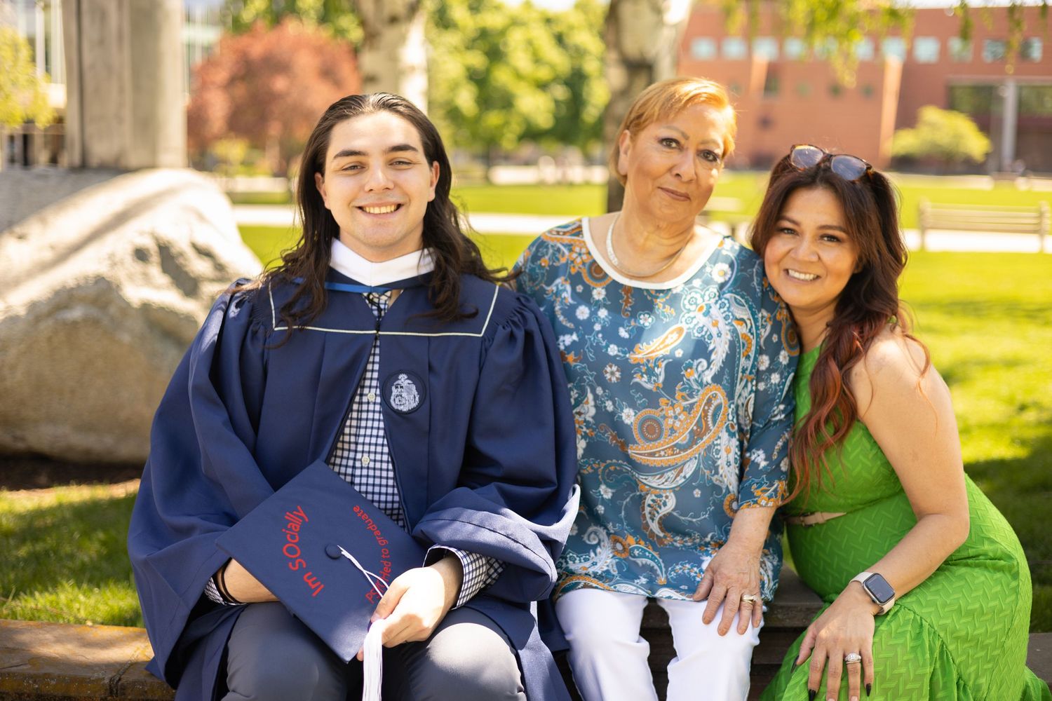 Group celebrates graduation on a sunny day in a park, with one wearing a navy graduation gown.