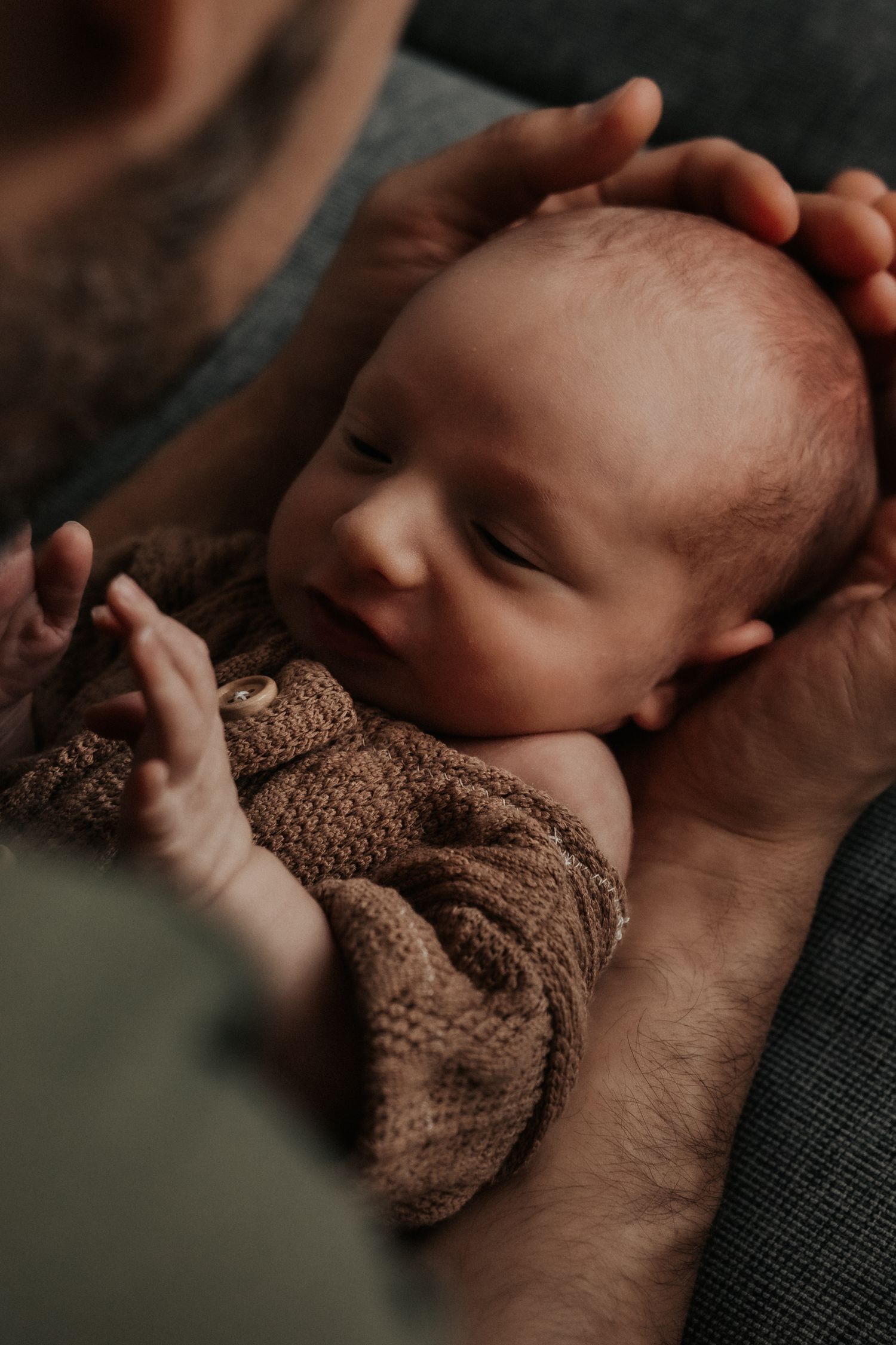 A sleeping baby wearing a brown knit sweater is cradled gently in caring hands.