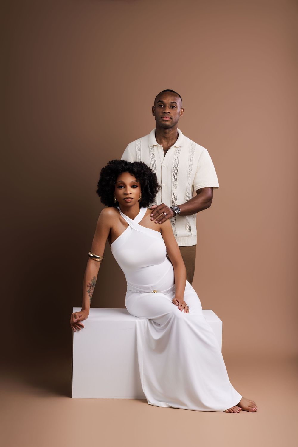 A pregnant Black woman in a white halter dress sits on a white cube beside a man in a cream shirt against a brown backdrop.
