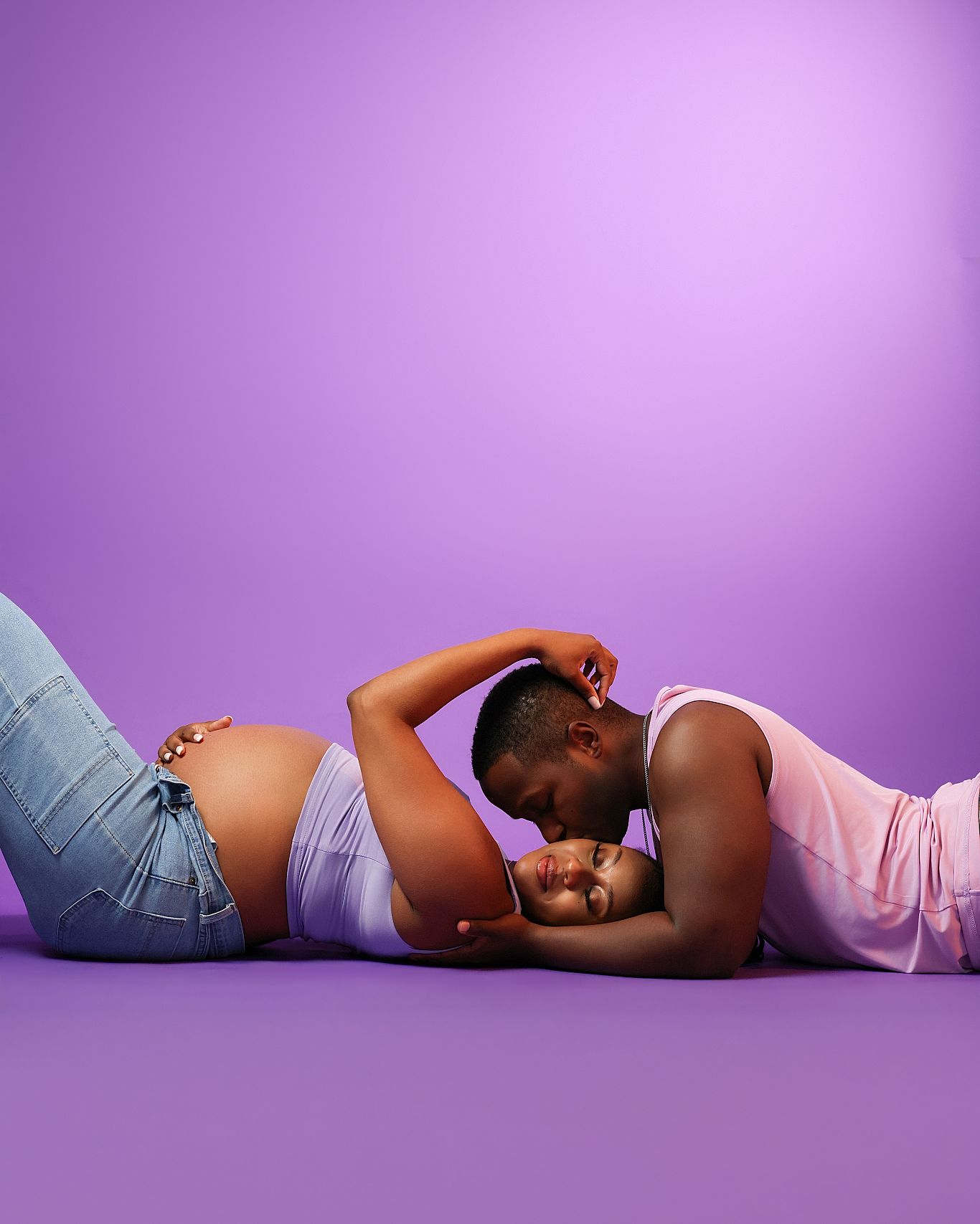 Pregnant Black woman and partner lying on purple studio floor, sharing tender moment with her baby bump visible.