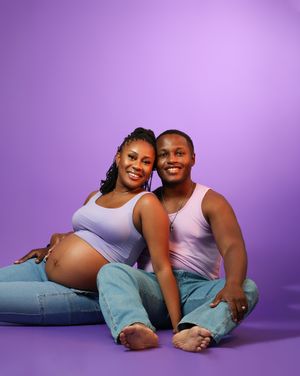 Smiling pregnant couple sitting on floor against purple background, woman in lavender tank top showing baby bump, man in pink tank top.