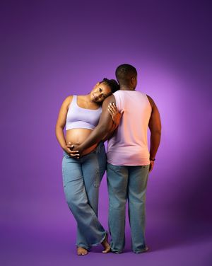 Pregnant woman in lavender crop top leans on partner against purple background, both wearing jeans.