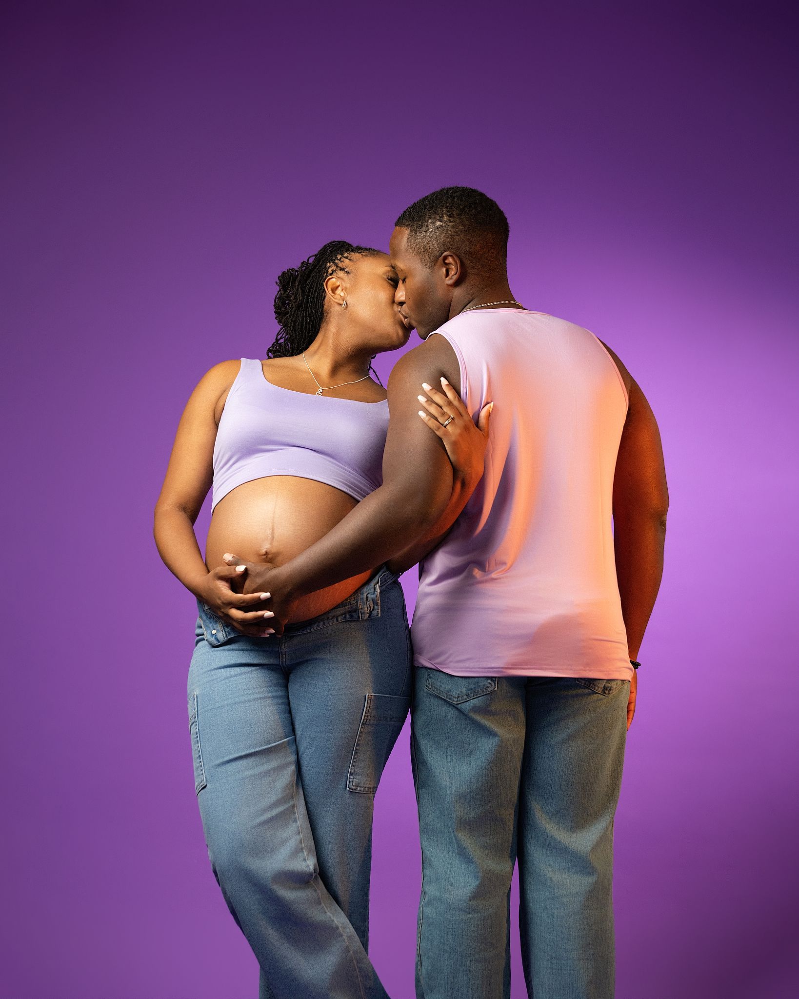 A pregnant Black woman in a lavender crop top and jeans kisses her partner against a purple studio background.