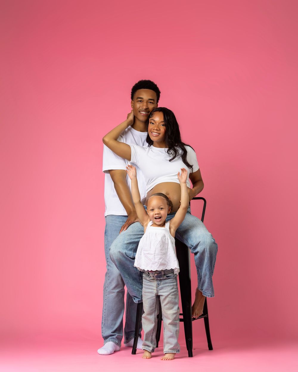 A happy family poses on a pink background, parents in white tees and jeans with their toddler daughter.